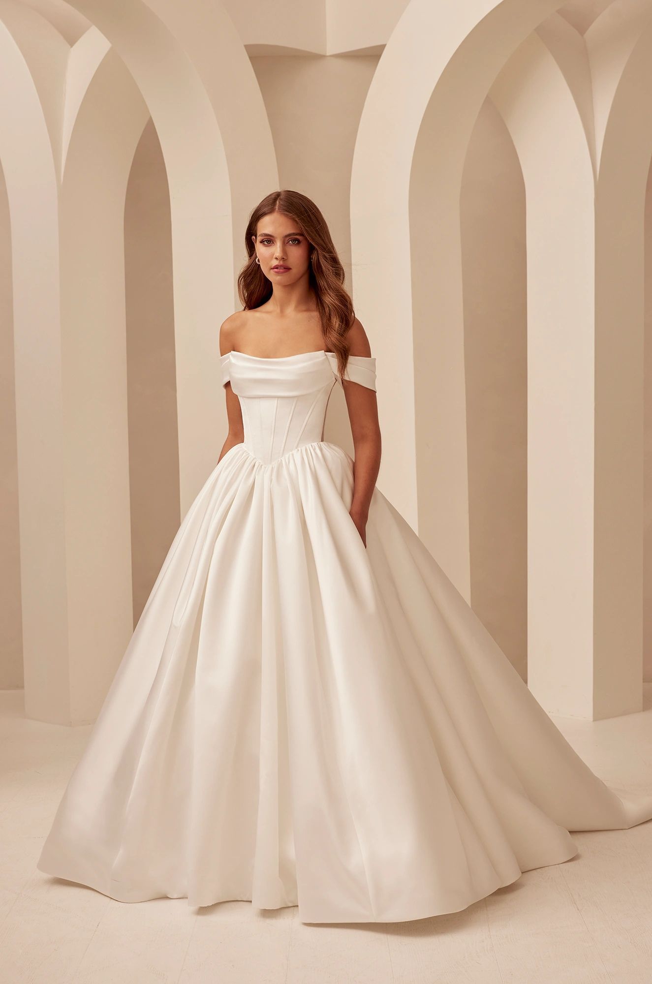 A woman wearing an off-the-shoulder, white satin ball gown with a draped bodice, standing in a room with arched pillars.
