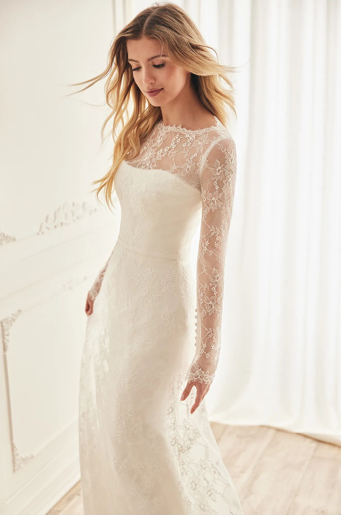 A person wearing a long-sleeved, white, lace wedding dress with an illusion neckline stands in a light-filled room.