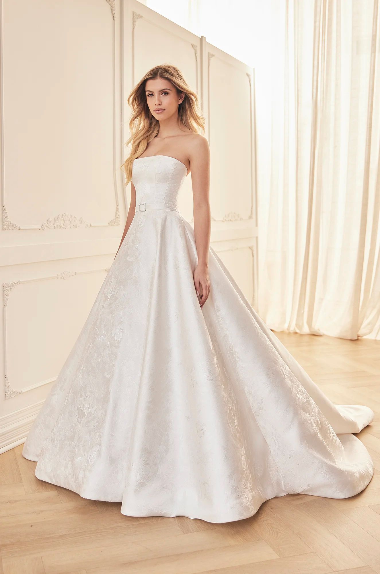 A strapless white ballgown with a textured, floral-patterned skirt stands in a bright, minimalist room with curtains.