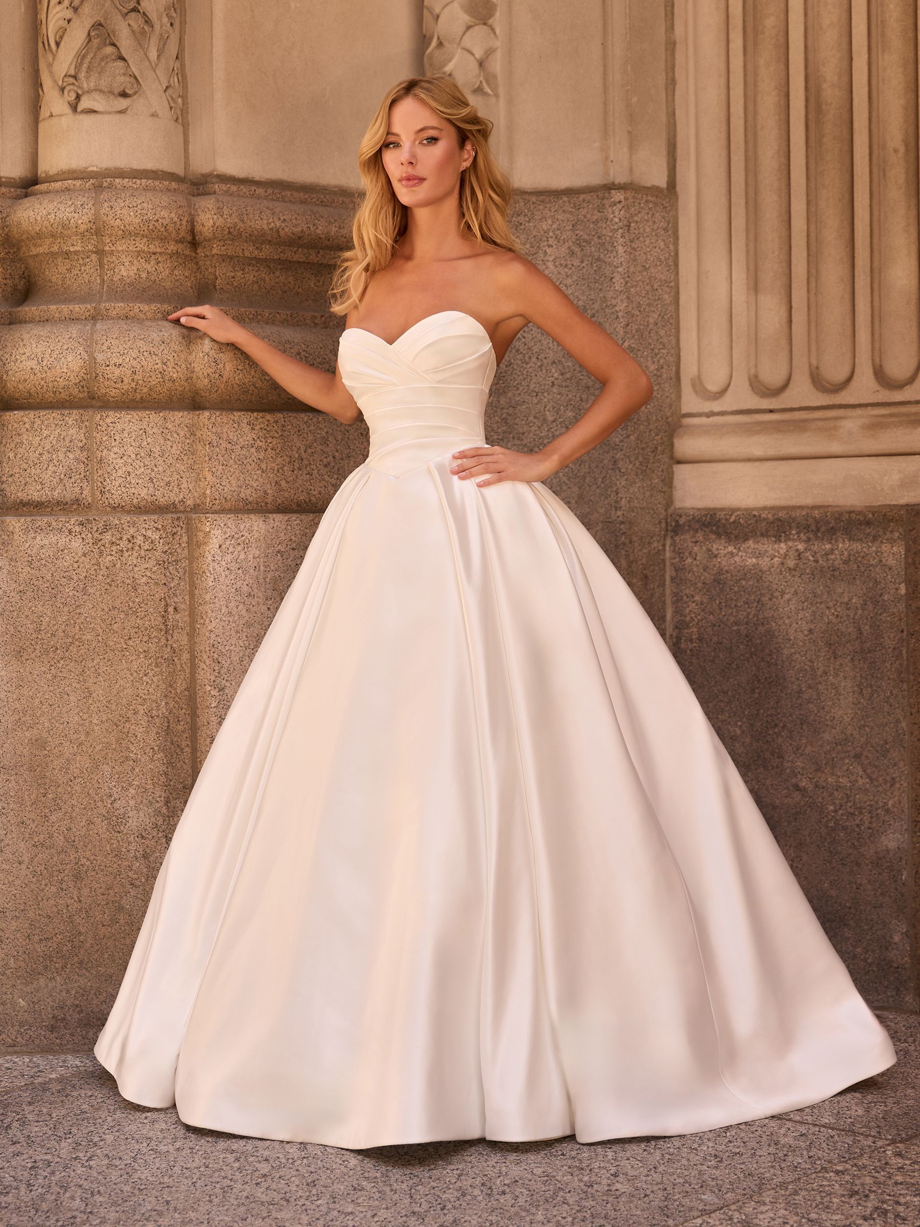 A person wears a strapless, floor-length white satin ballgown with a sweetheart neckline, standing by stone pillars.