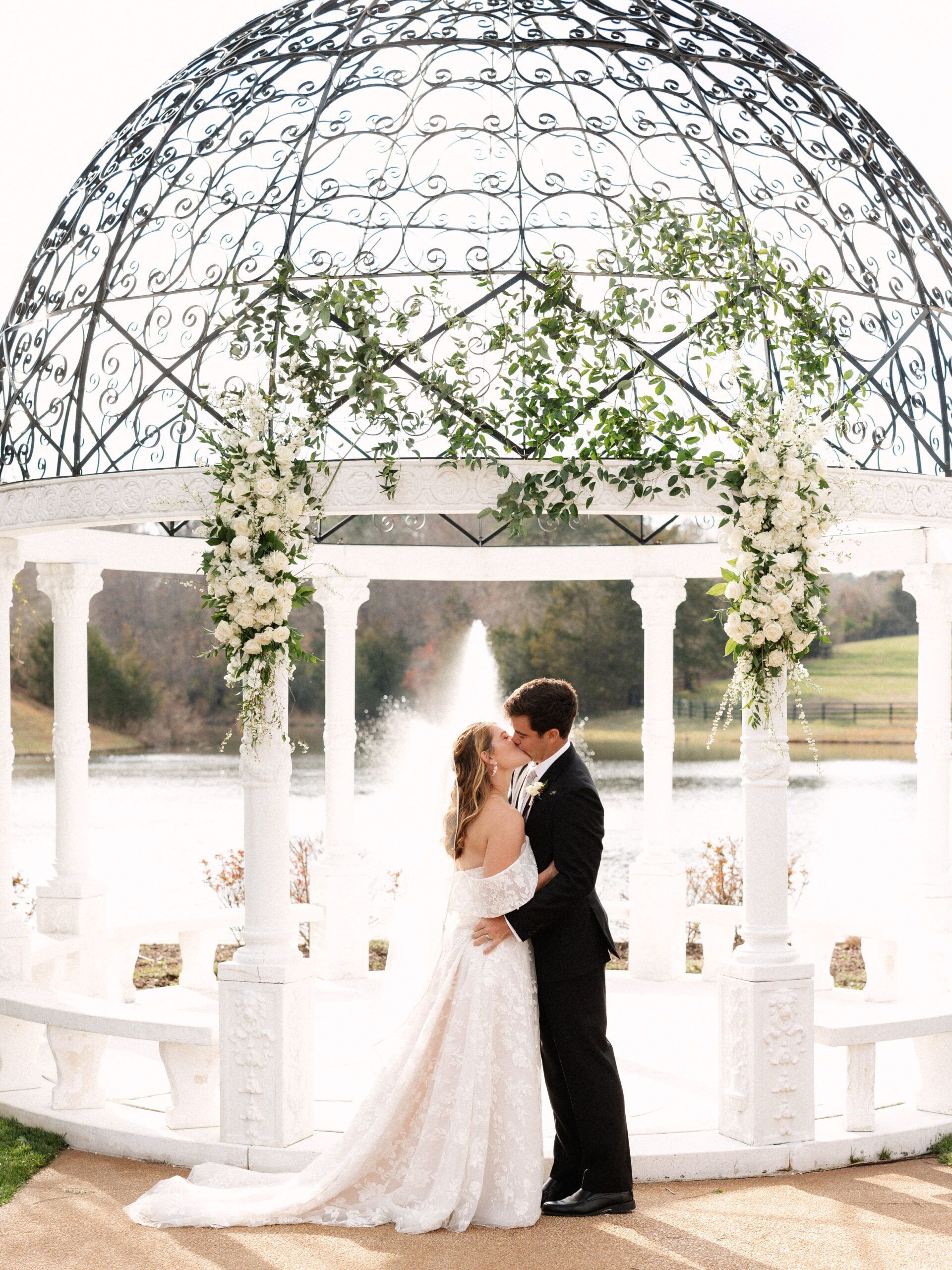 A couple kisses under a white, flower-adorned gazebo overlooking a pond with a fountain in the background.