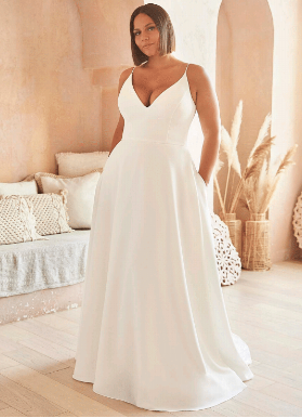 A person wears a minimalist white wedding dress with a V-neck and pockets in a bright, neutral-toned room.