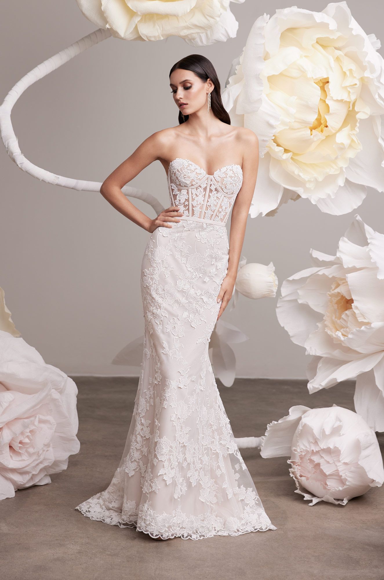 A model poses in a strapless, floor-length lace wedding dress against a backdrop of large, cream-colored paper flowers.