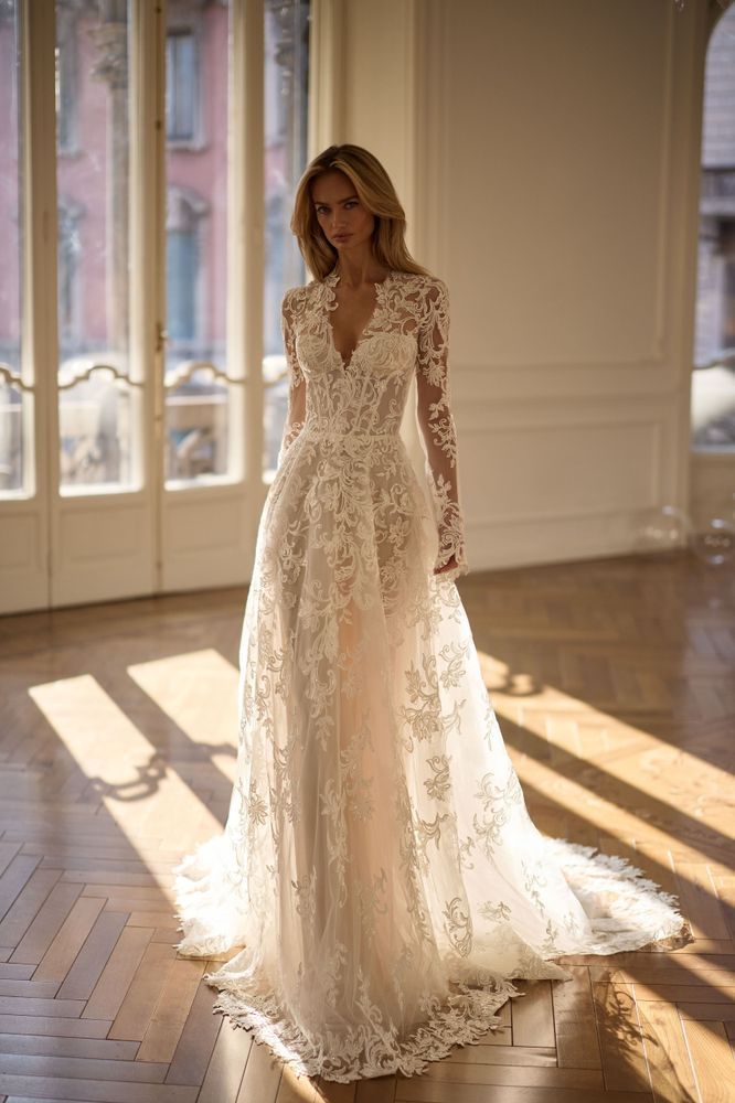 A person wearing a long-sleeved, floor-length white lace wedding gown with a deep V-neck in a sunlit room.
