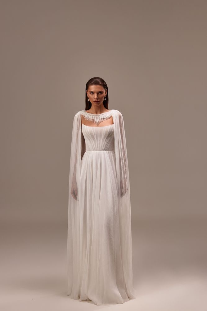 A person wearing a long, white, pleated bridal gown with a sheer cape overlay against a minimalist neutral background.