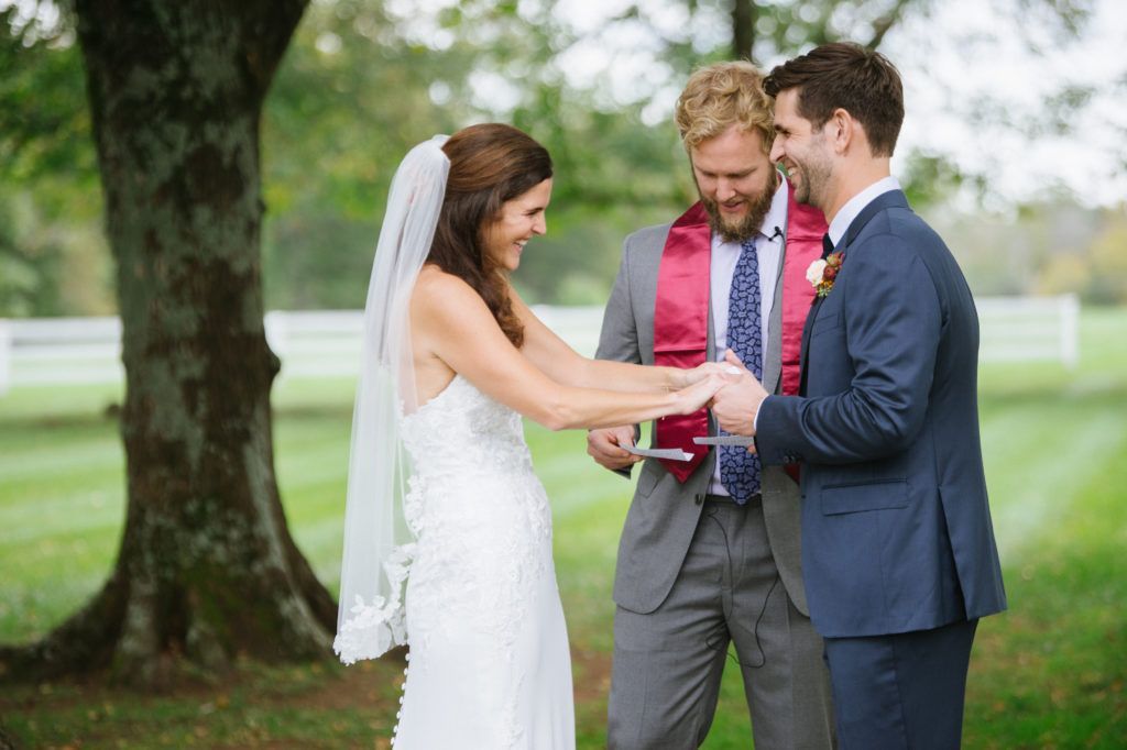 A bride and groom holding hands and laughing during their outdoor wedding ceremony, with an officiant standing nearby.