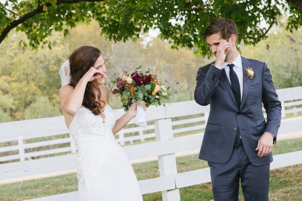 A bride and groom standing in front of a white fence, both wiping away tears of joy while the bride holds a bouquet.