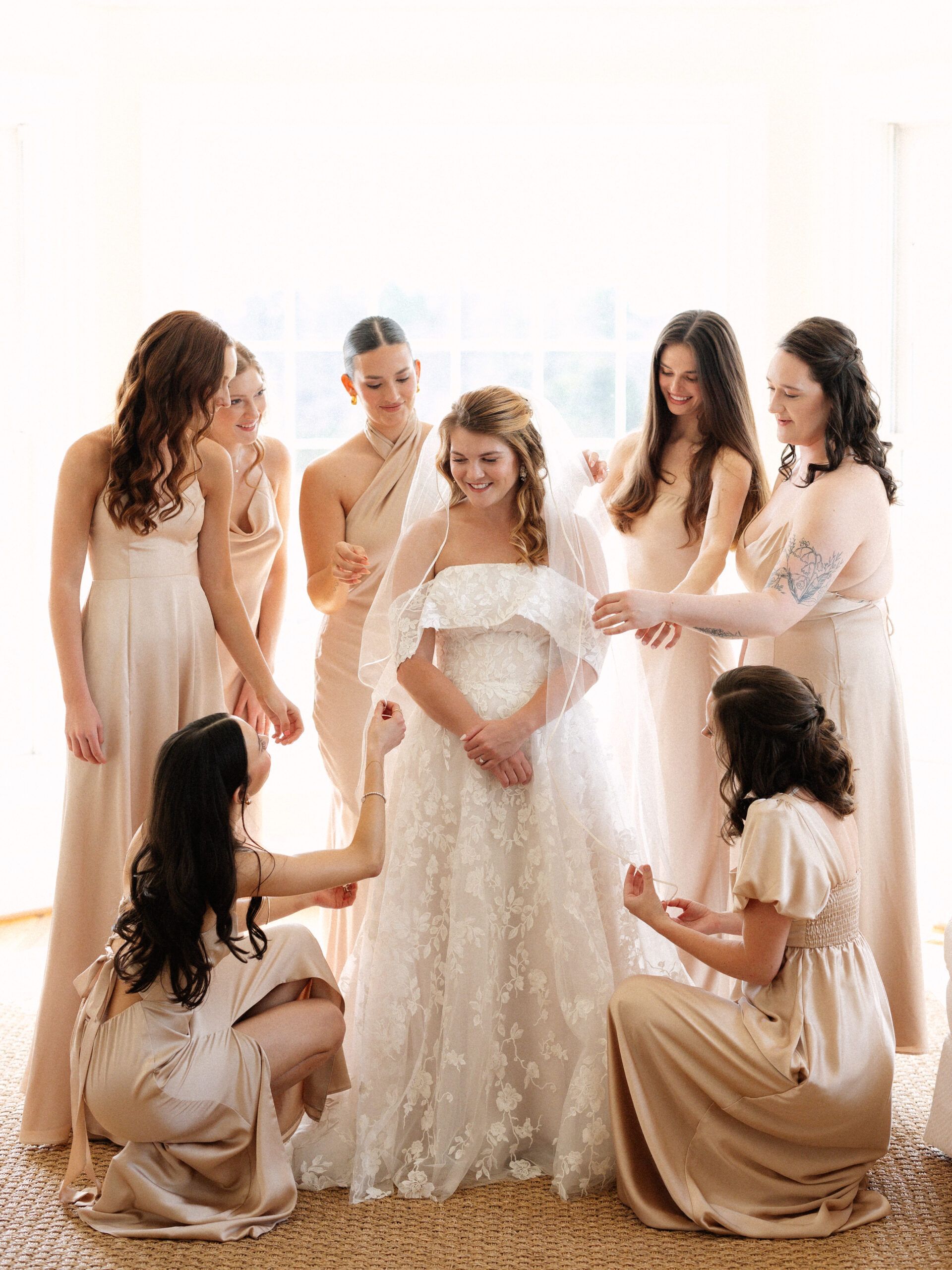 A bride in a white lace gown stands surrounded by seven people in matching champagne-colored dresses, helping her get ready.