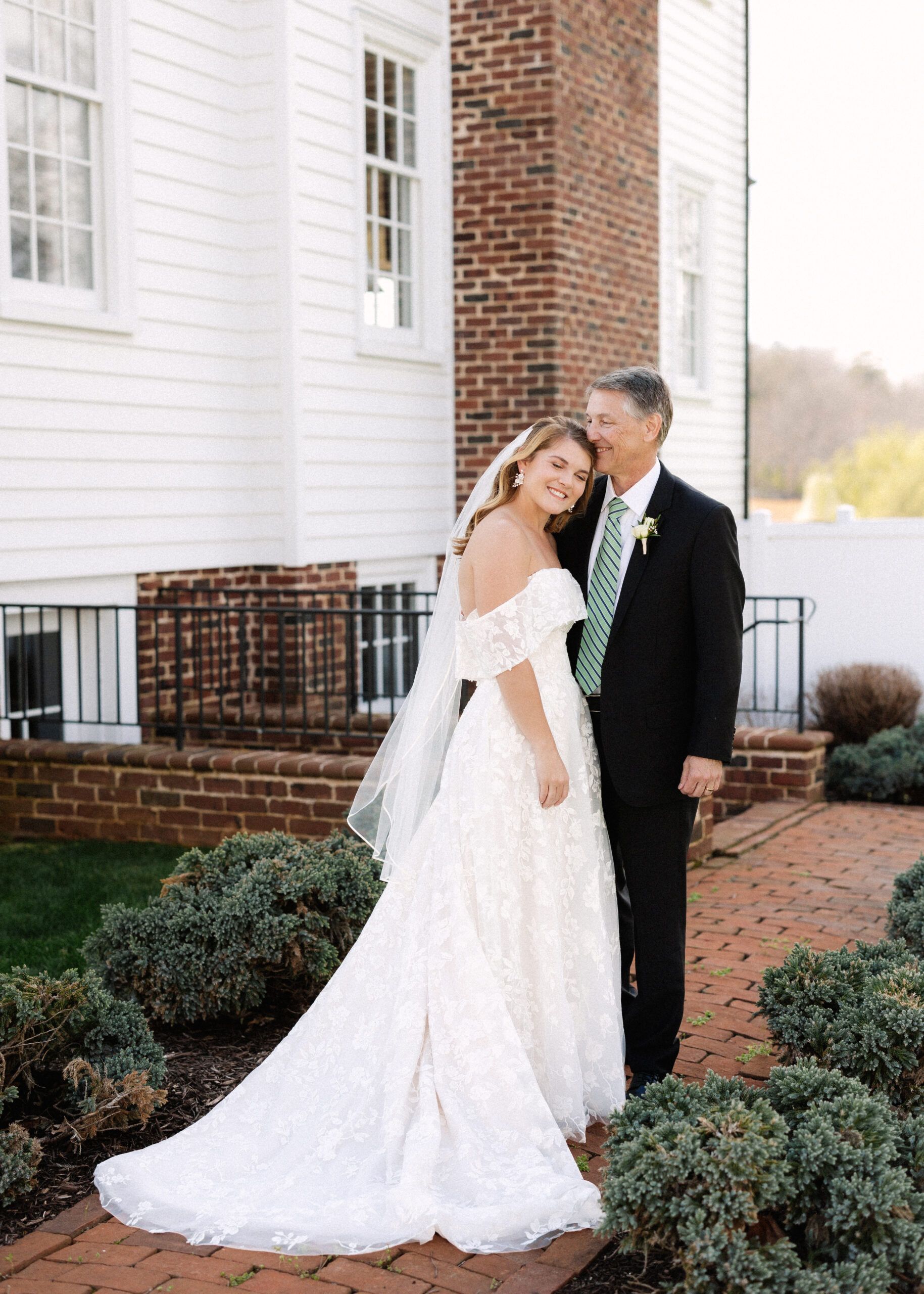 A bride in a white lace dress and veil standing beside a person in a suit on a brick path by a white house.