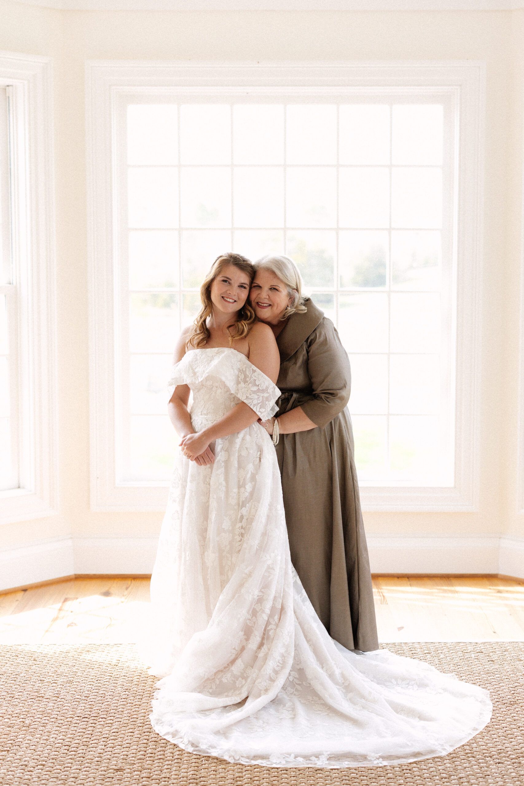 A bride in a white lace wedding dress stands by a window, held from behind by a person in a long brown dress.