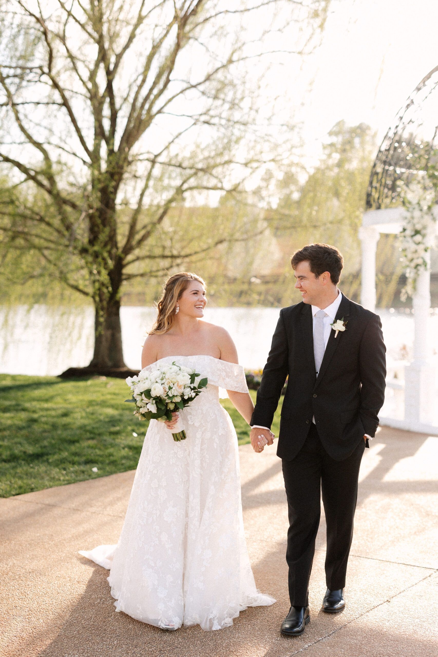 A bride and groom holding hands and smiling at each other while walking outdoors near a lake and a large tree.