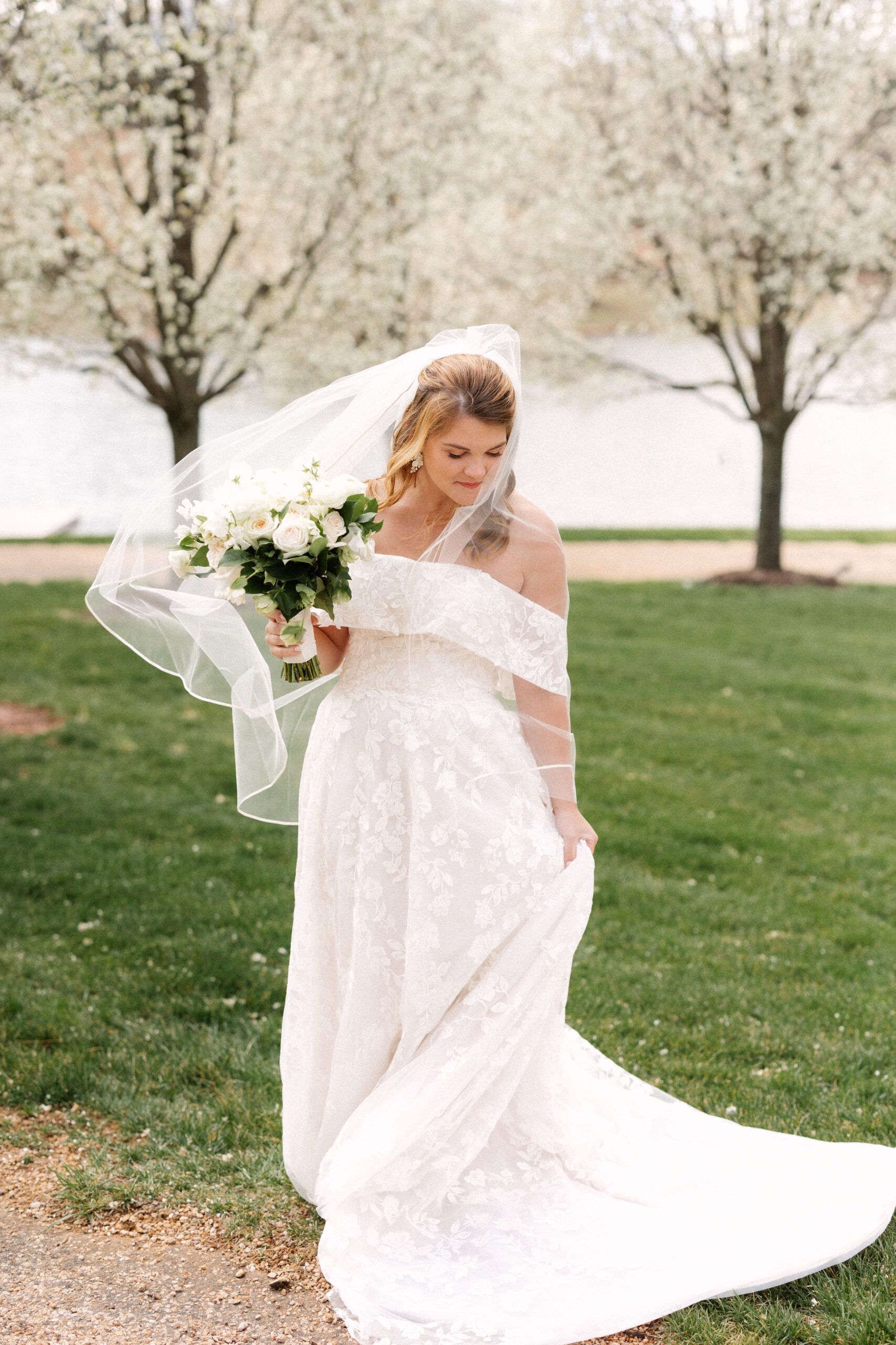 A bride in a lace off-the-shoulder gown holds a bouquet outdoors as her veil blows in the wind before blooming trees.