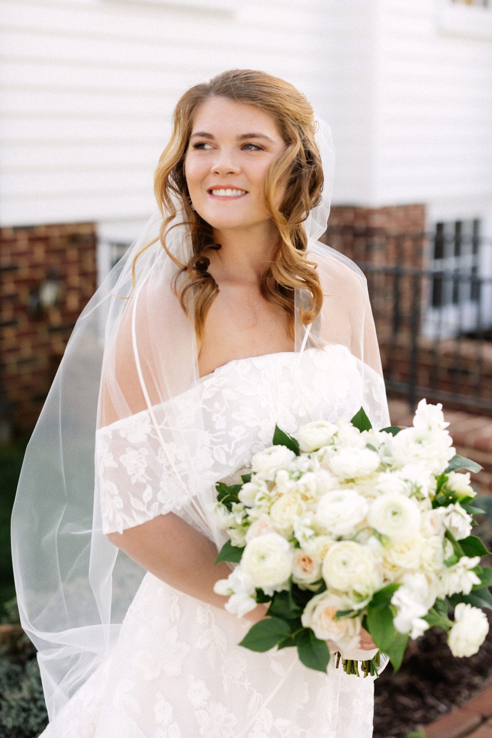 A smiling bride wearing an off-the-shoulder lace dress and veil, holding a bouquet of white roses outdoors.