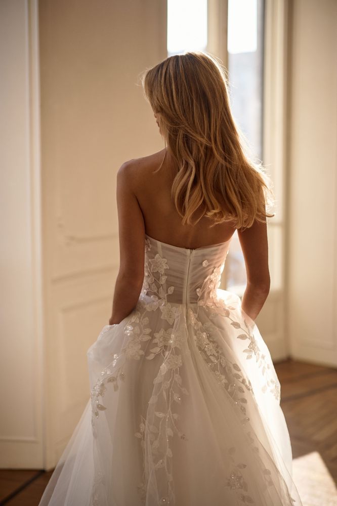 A person wearing a strapless white wedding dress with floral embroidery, standing in a sunlit room.