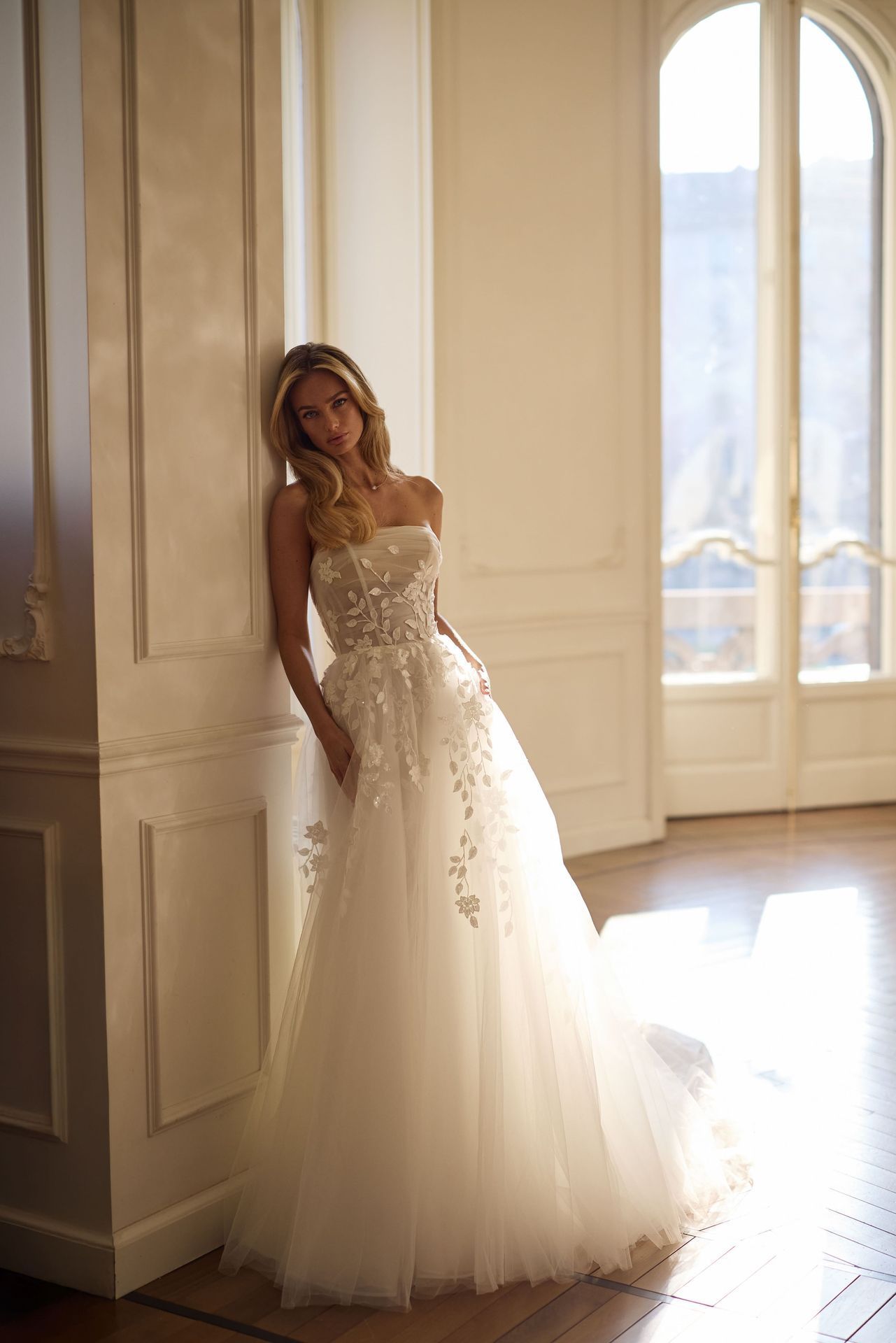 A person stands leaning against a wall in a sunlit room, wearing a strapless, floral-appliquéd white A-line wedding dress.