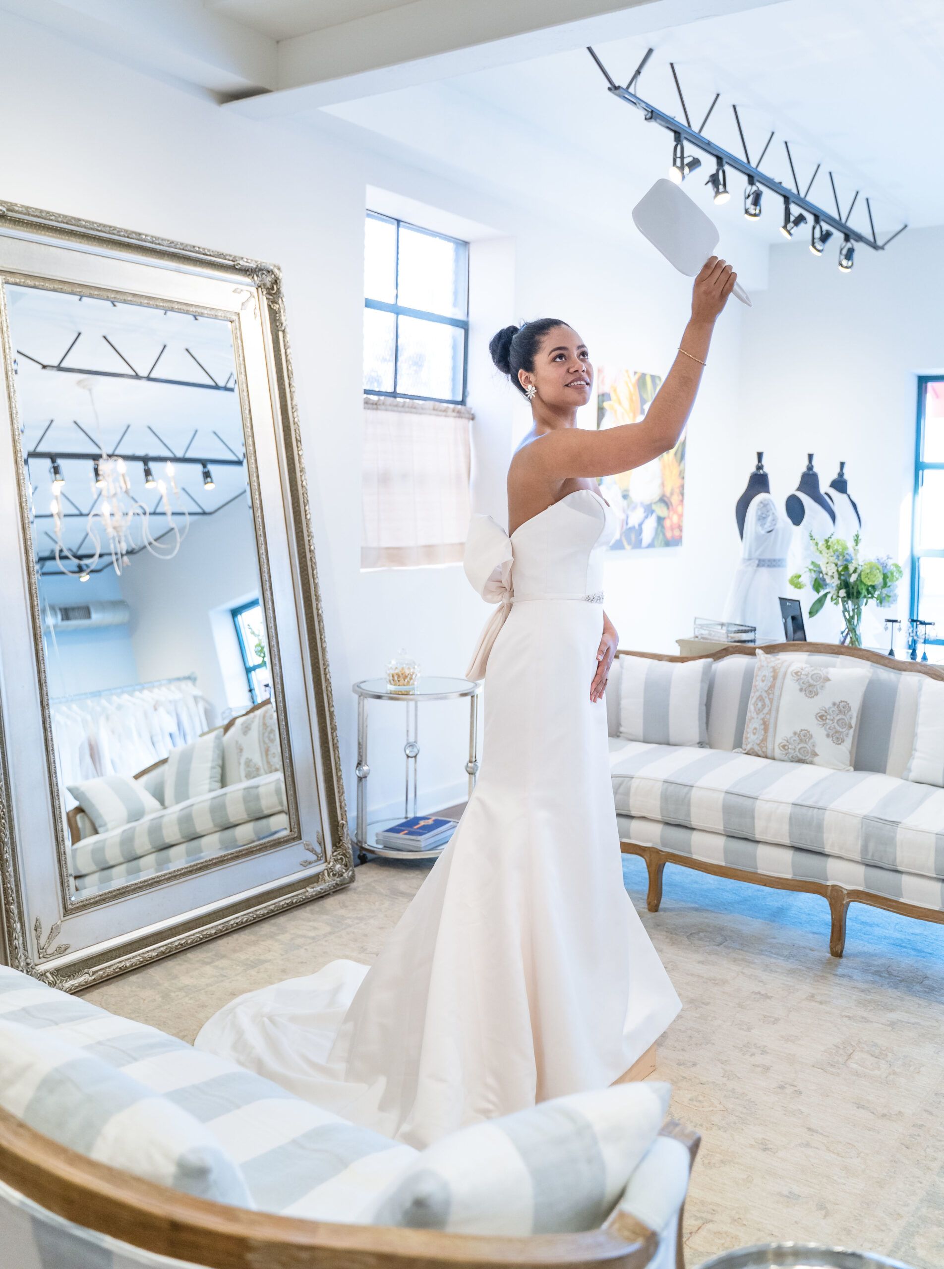 A person in a white wedding gown holding a mirror above their head in a boutique with mirrors and striped furniture.