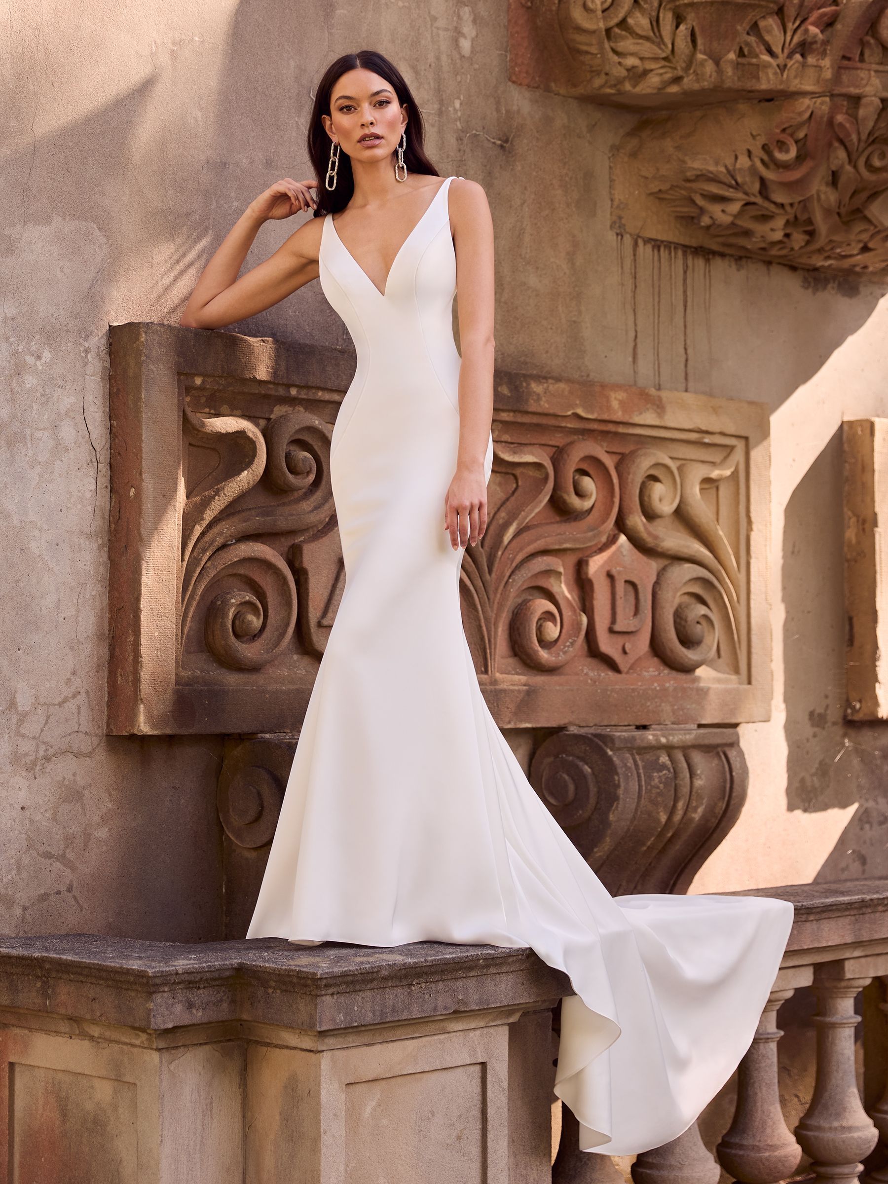 A person wears a form-fitting, sleeveless white V-neck bridal gown with a train, standing by an ornate stone structure.