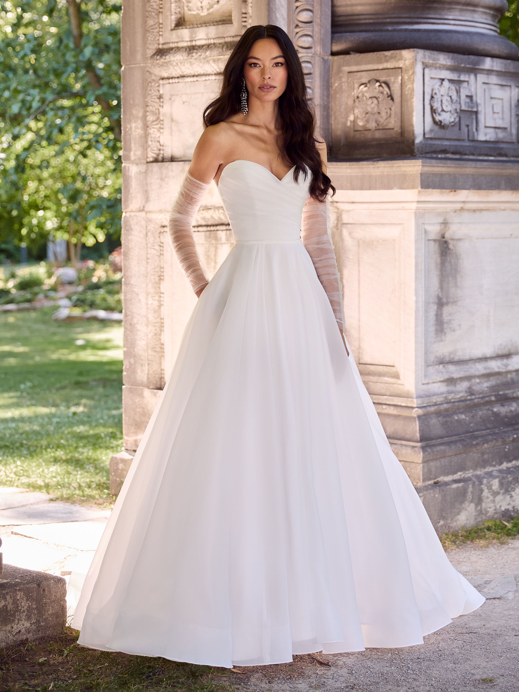 A person wearing a strapless white A-line wedding dress with lace sleeves, standing in a garden near a stone pillar.