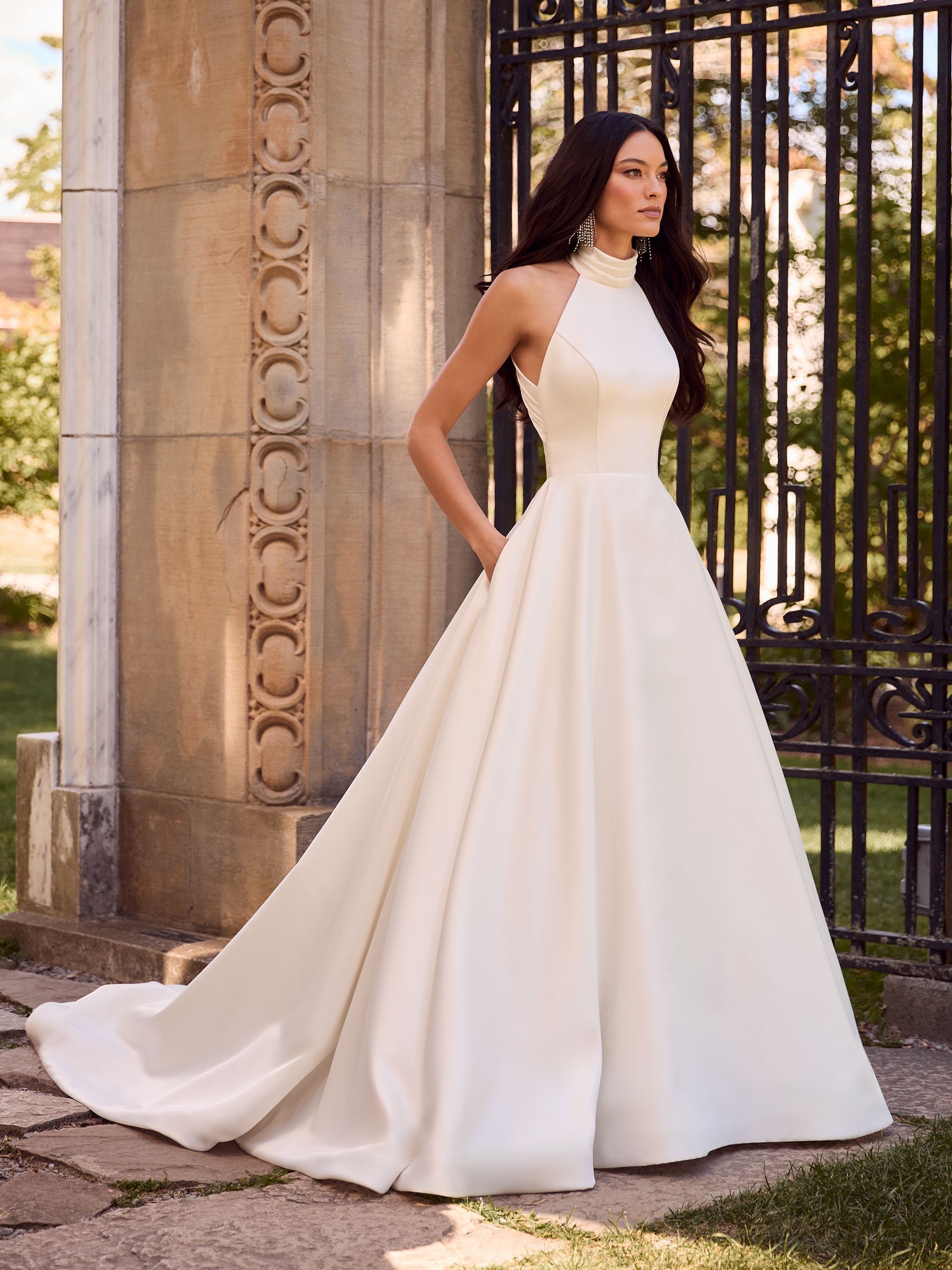 A person stands outdoors wearing a floor-length, ivory-colored, high-neck wedding gown with a full skirt and pockets.