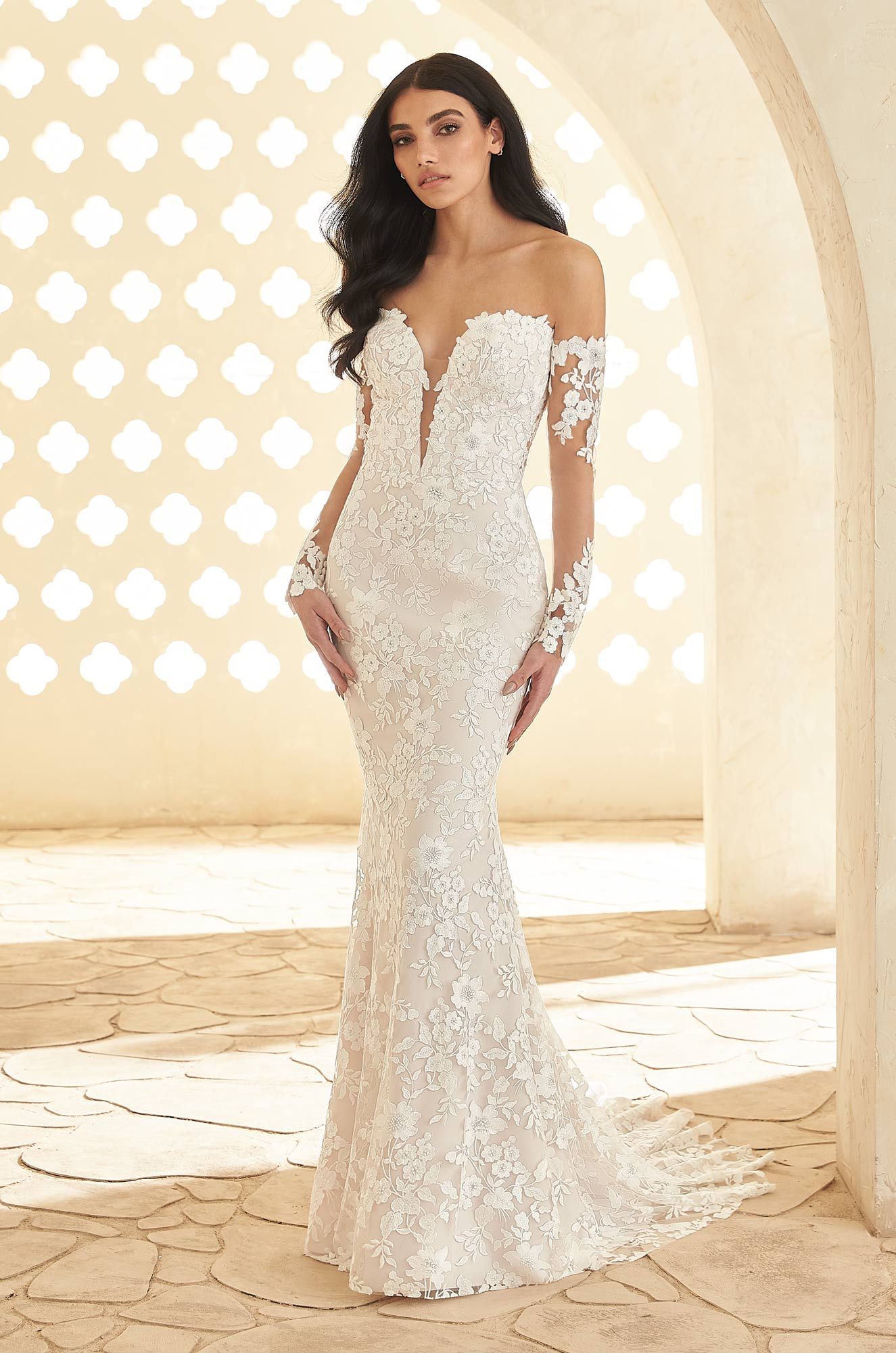 A model wears a form-fitting, off-the-shoulder lace wedding dress with long sleeves in a sunlit, arched stone setting.
