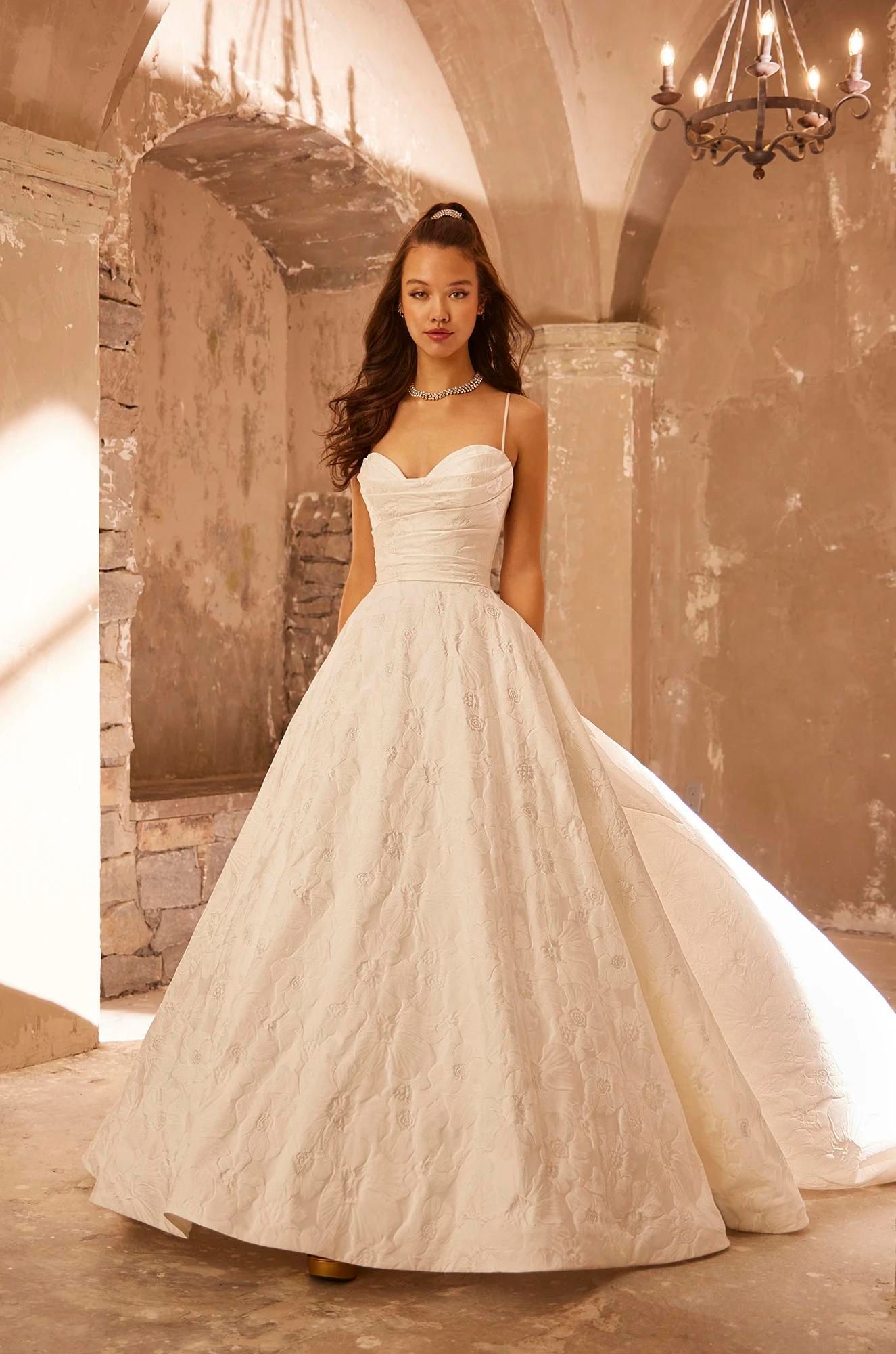 A person in a floor-length white ballgown with a sweetheart neckline and textured fabric, posing in a stone-arch corridor.