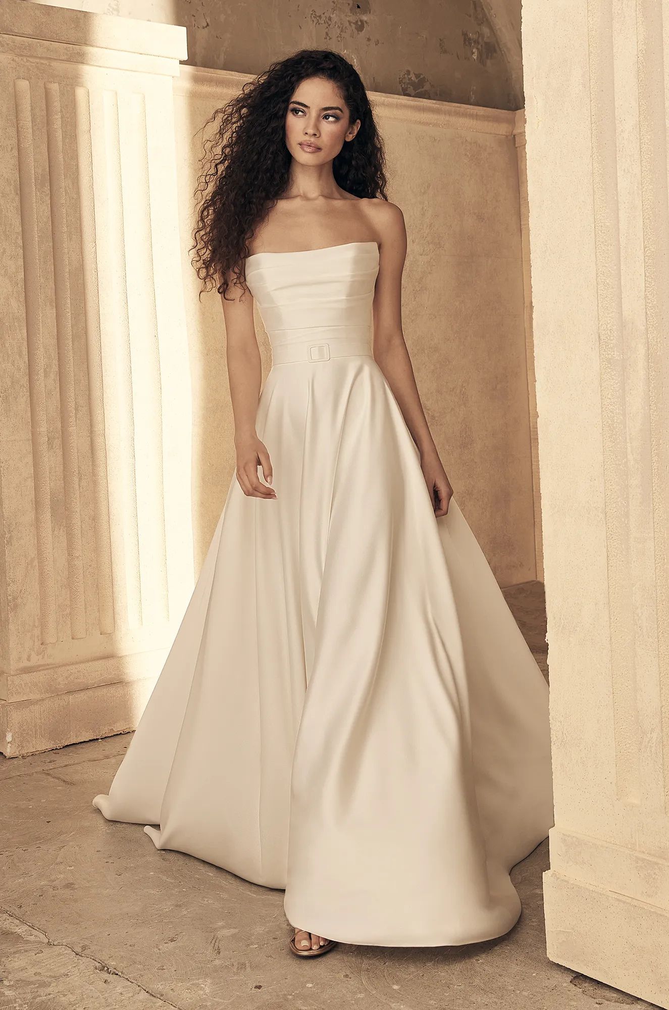 A person with long, curly dark hair wearing a floor-length, strapless cream-colored gown, posing in a stone-columned hall.