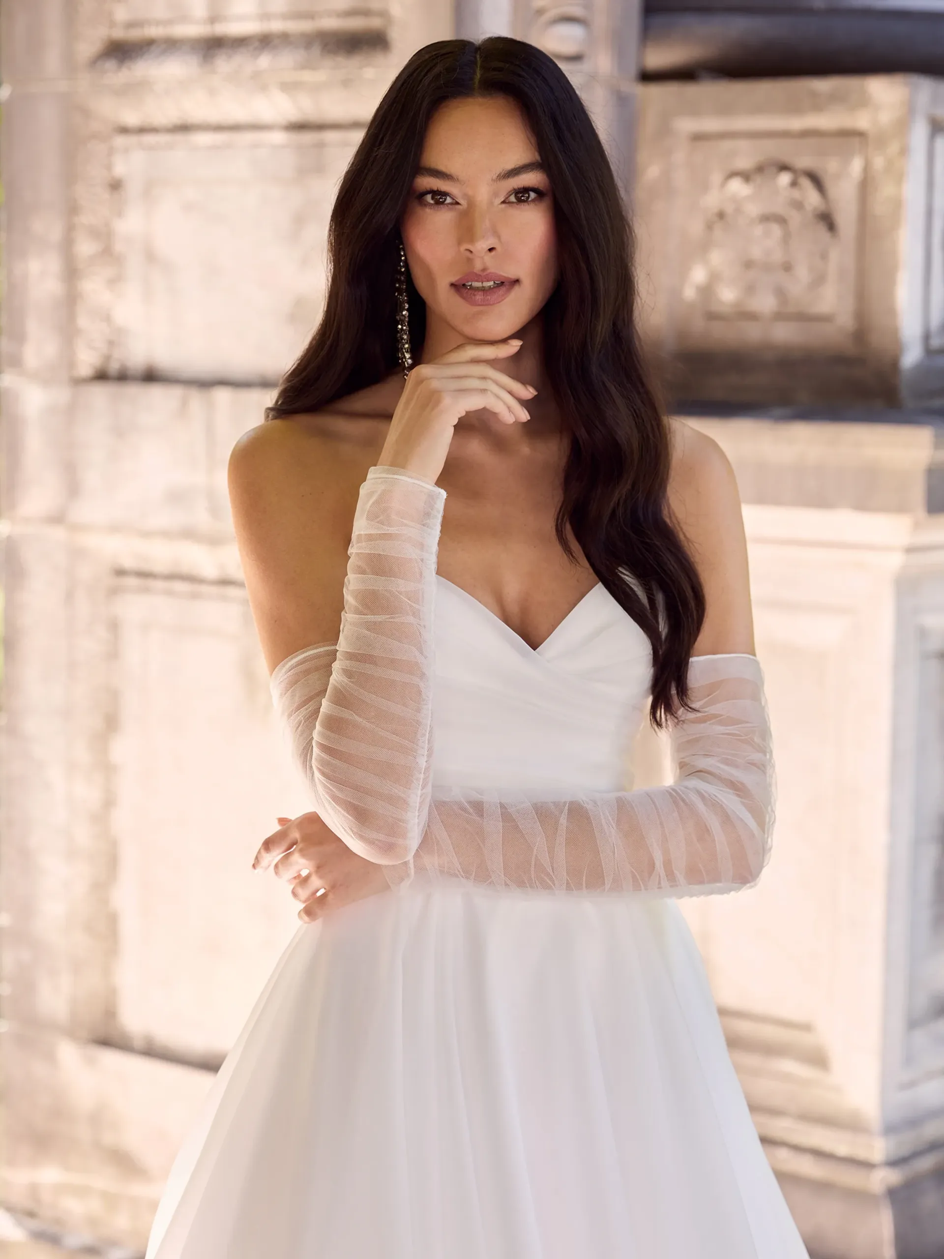 A person with long dark hair wears a white strapless wedding gown with matching sheer, beaded long gloves.