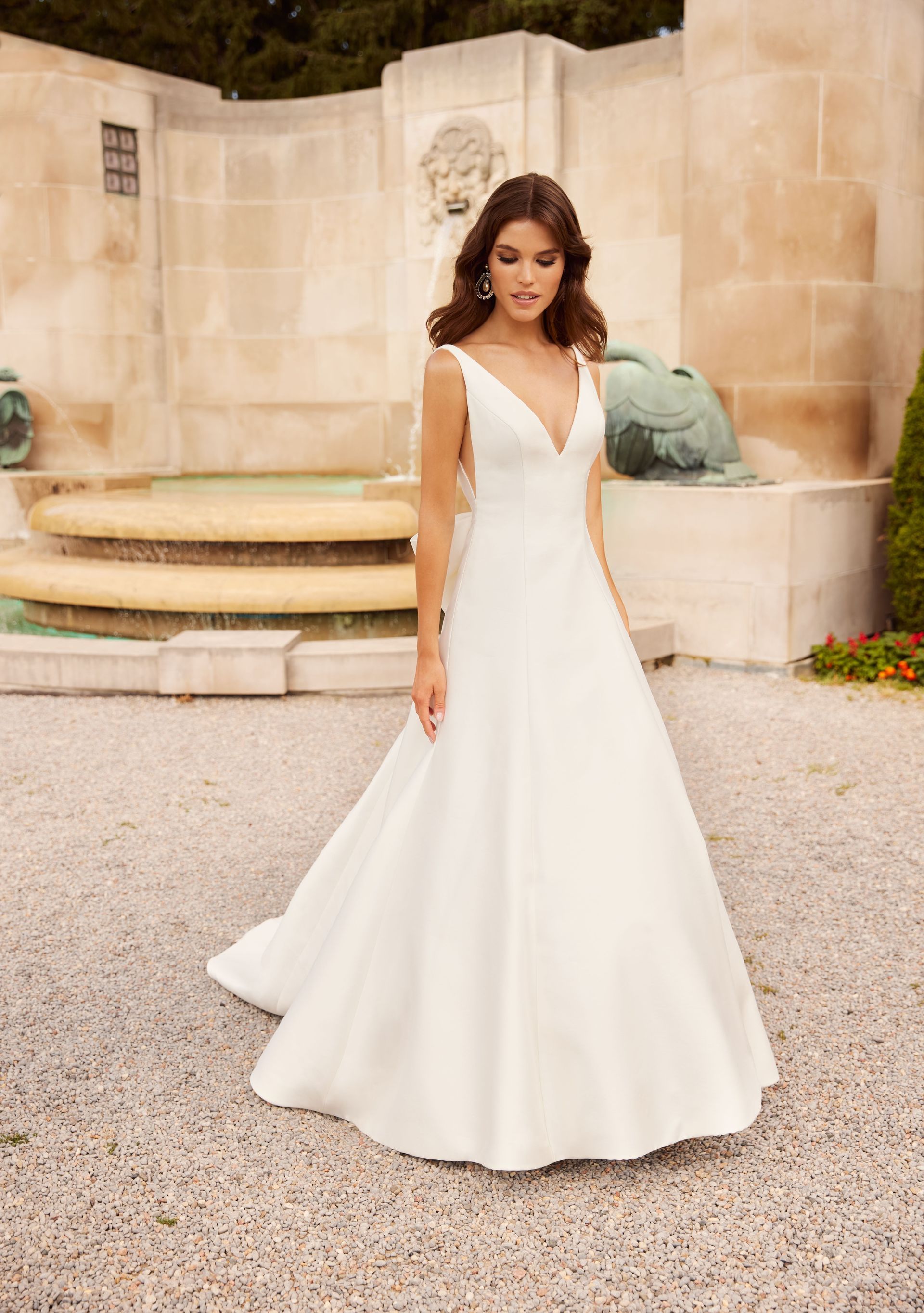 A person in a floor-length white A-line wedding dress with a V-neckline, standing outdoors in front of a stone fountain.