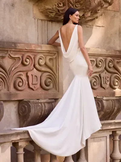 A person in a form-fitting, white bridal gown with a low cowl back and train, posing against a stone architectural wall.