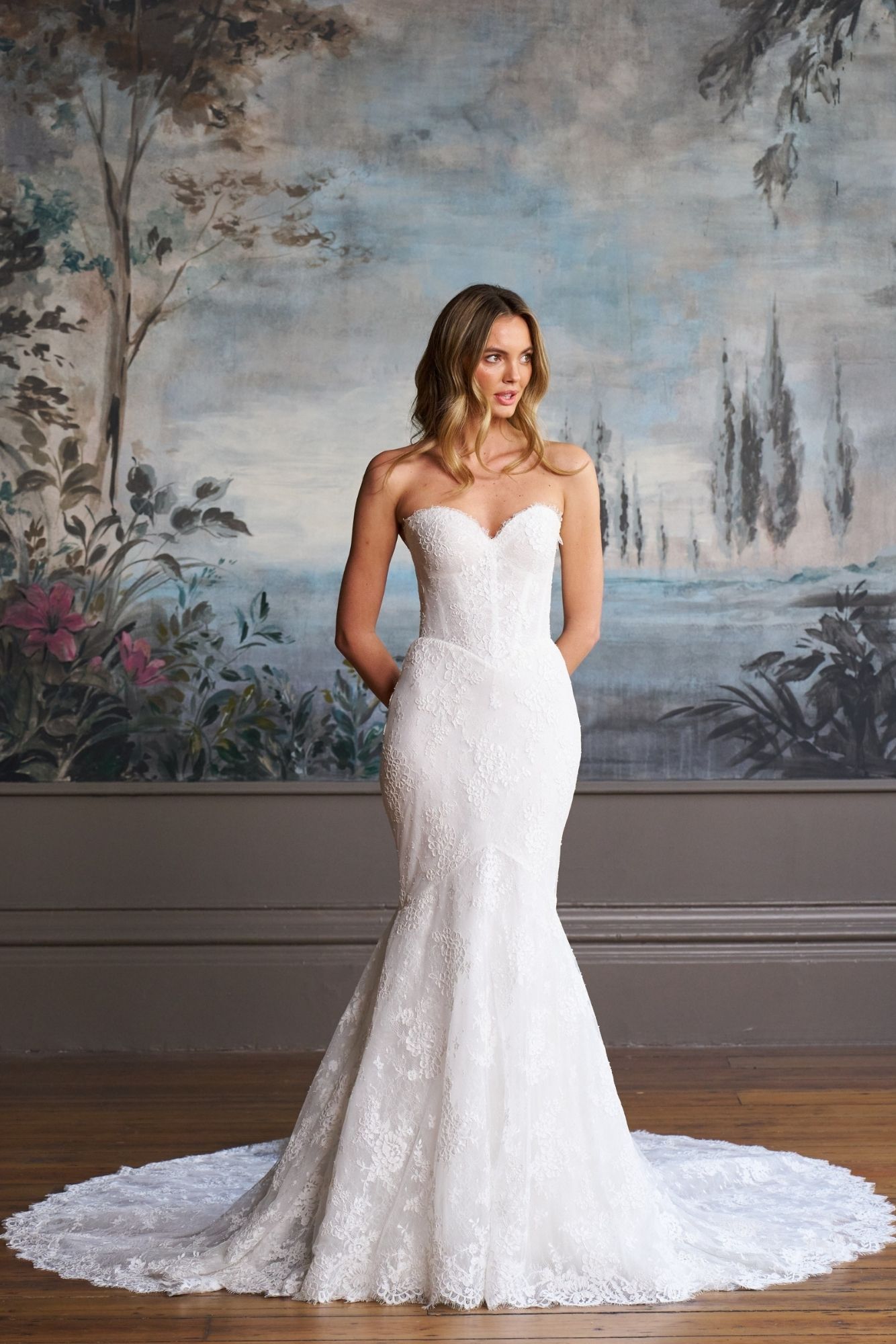 A person wearing a white, strapless, lace mermaid-style wedding dress standing in front of a mural background.