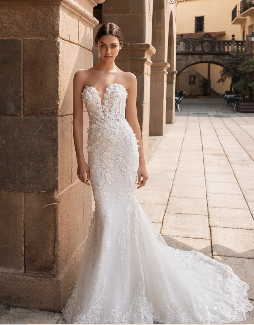 A person wearing a strapless, floor-length white mermaid-style wedding dress with lace details, standing outdoors.