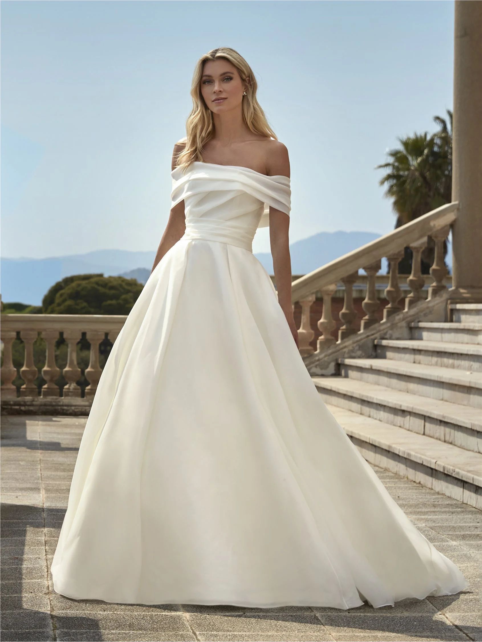A model wearing a white, off-the-shoulder A-line wedding dress with a full skirt, standing on stone stairs outdoors.