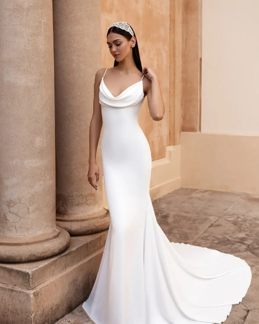 A person wearing a white, floor-length, cowl-neck satin wedding dress with a train, standing by stone pillars.