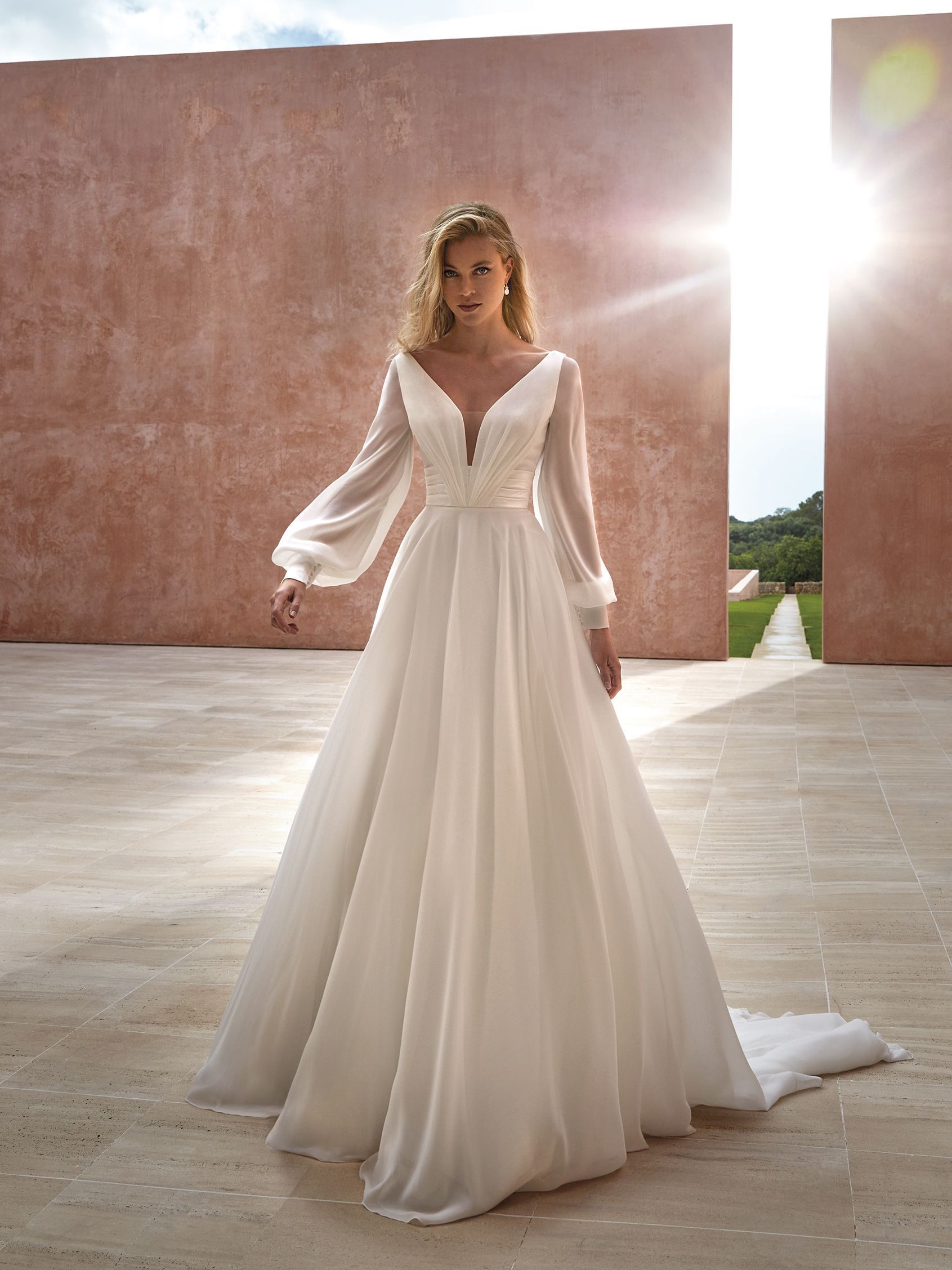 A person wearing a long-sleeved, white flowy wedding dress standing in front of large, sunlit stone walls.