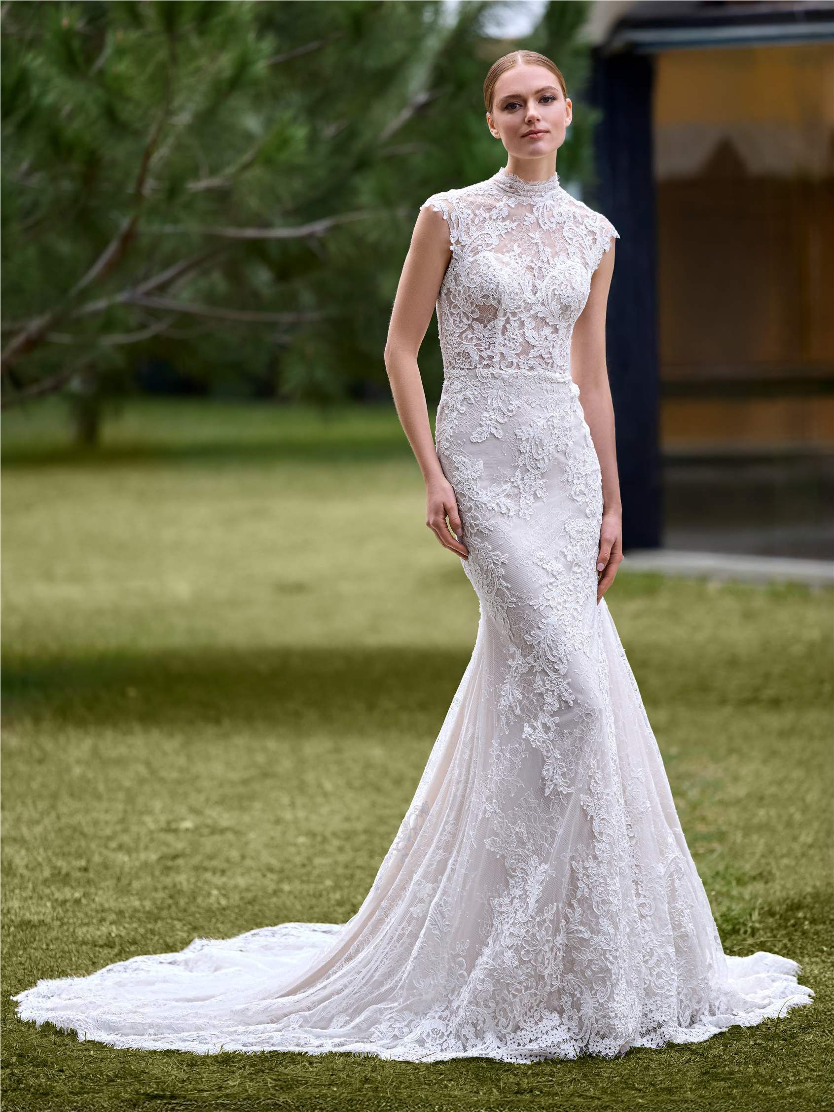 A model wearing a white, sleeveless, high-neck lace mermaid wedding dress standing on grass outdoors.