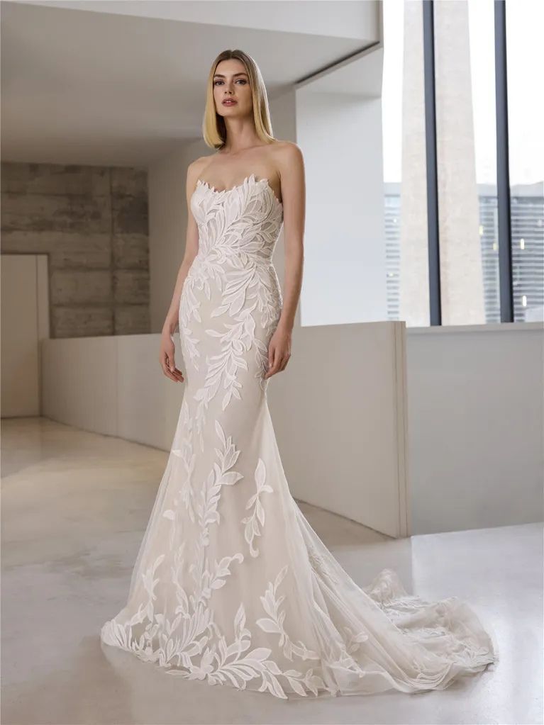 A floor-length, strapless, ivory bridal gown with intricate lace floral detailing and a small train, worn by a model.