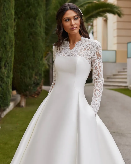 A person wears a white satin A-line wedding dress with a long-sleeved lace bodice, standing outdoors near trees.