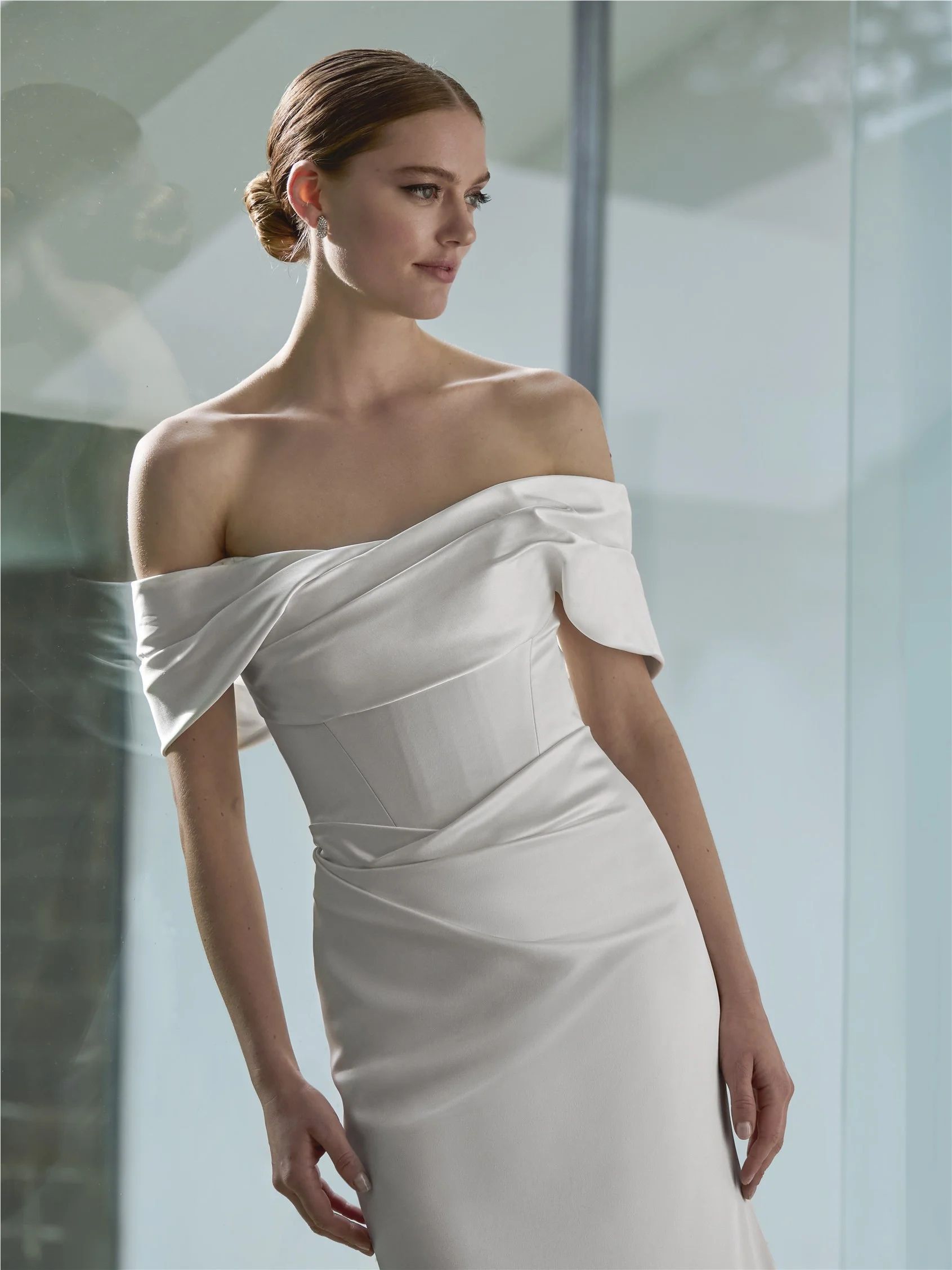 A model wearing a white off-the-shoulder satin gown with draped fabric detail on the bodice, standing against a glass wall.