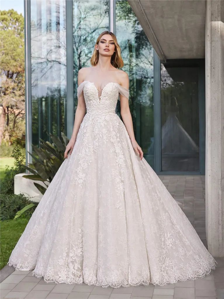 A model wears an off-the-shoulder, floral lace ballgown with a sweetheart neckline, standing in a modern outdoor setting.