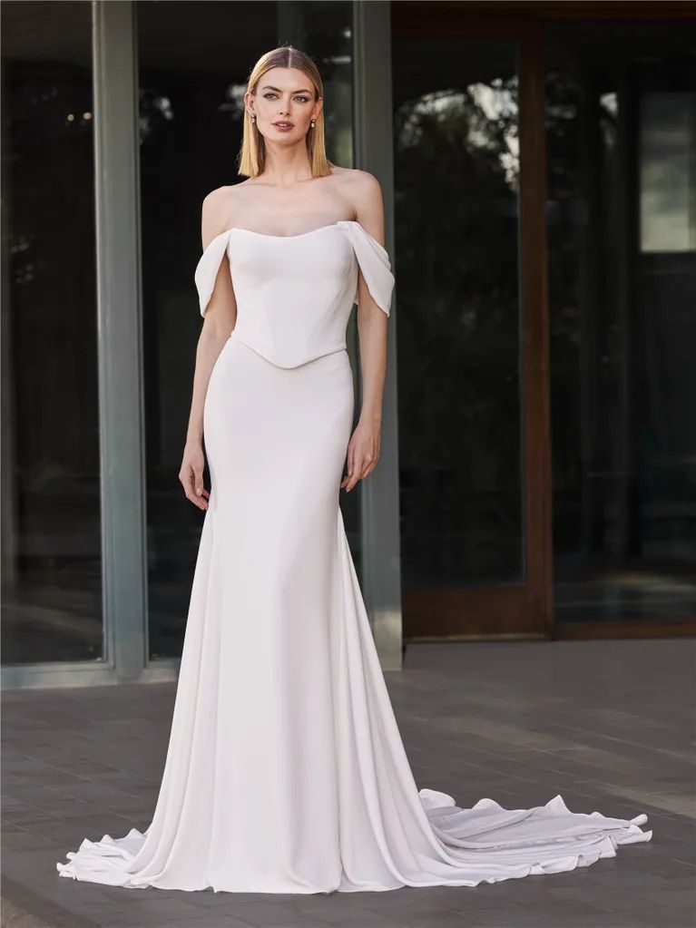A model wears a sleek, floor-length off-the-shoulder white wedding gown with a scalloped neckline and a small train.