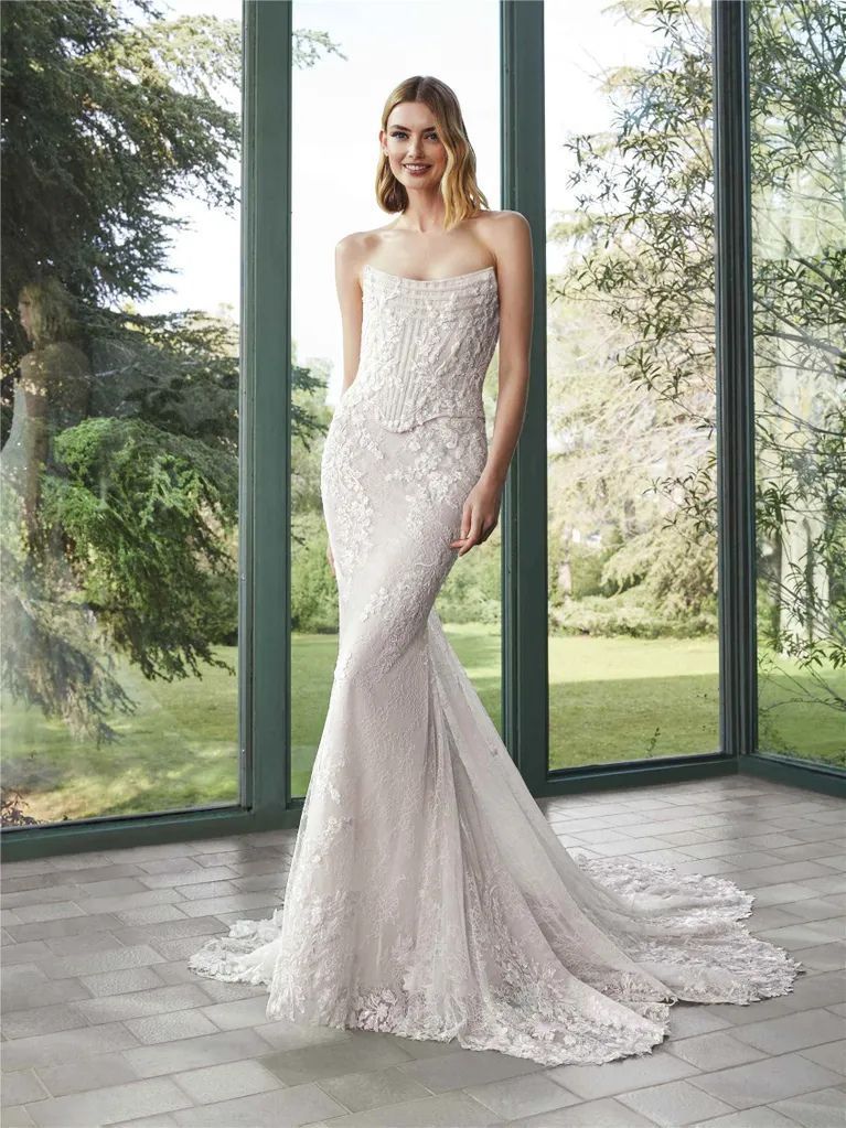 A person wearing a strapless, floor-length, textured white mermaid-style wedding dress standing in a sunlit conservatory.