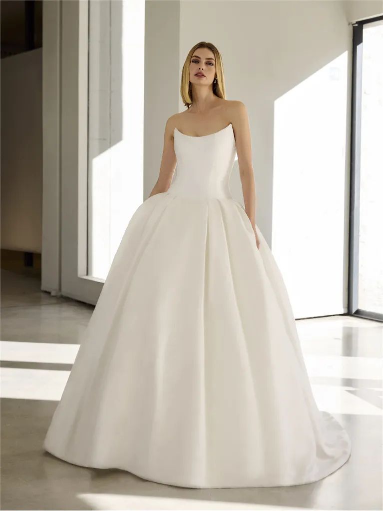 A person wears a strapless, white, floor-length ballgown with a structured bodice in a brightly lit, modern room.