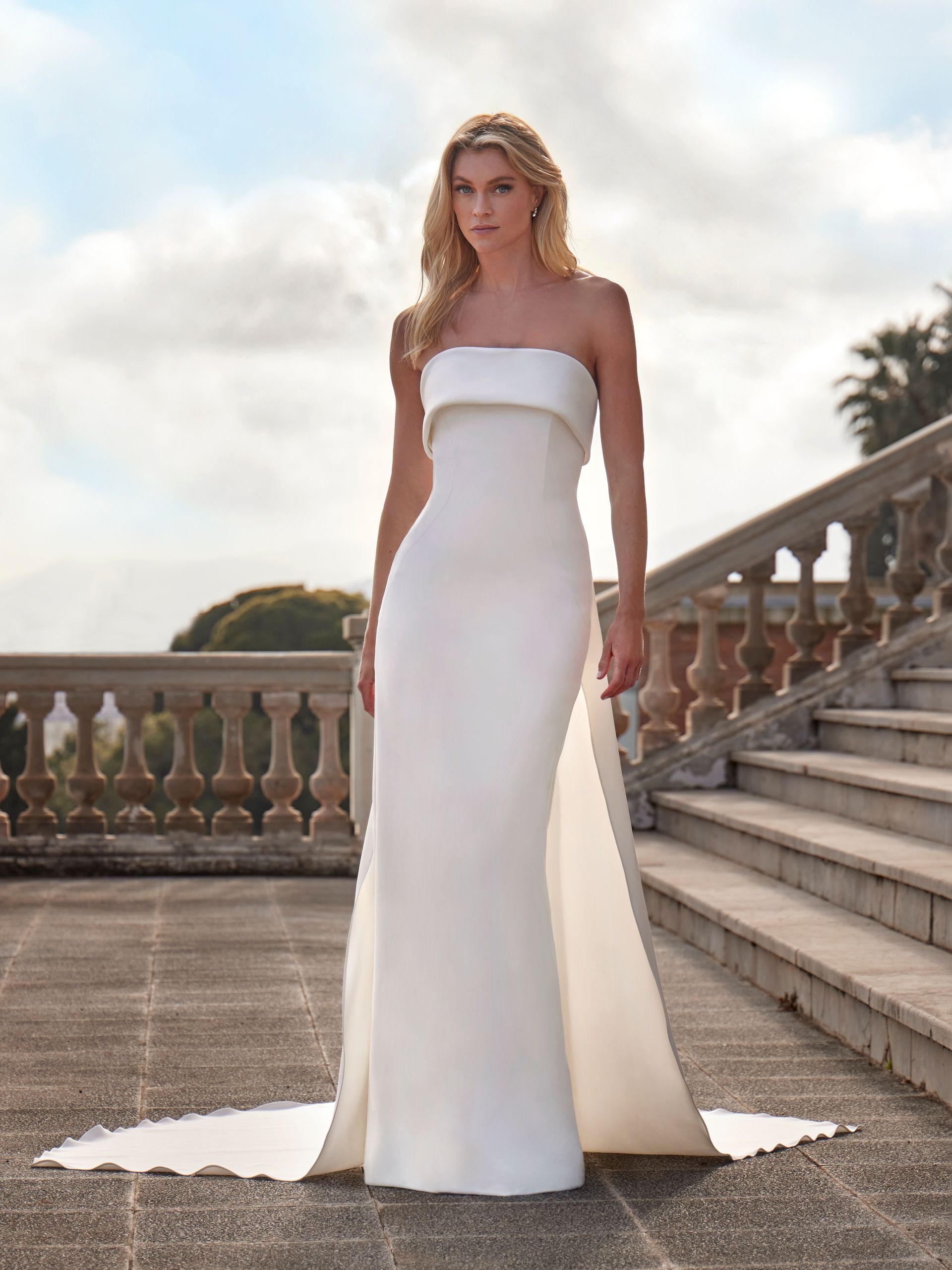 A person stands on stone steps outdoors wearing a white strapless floor-length wedding gown with a flowing train.