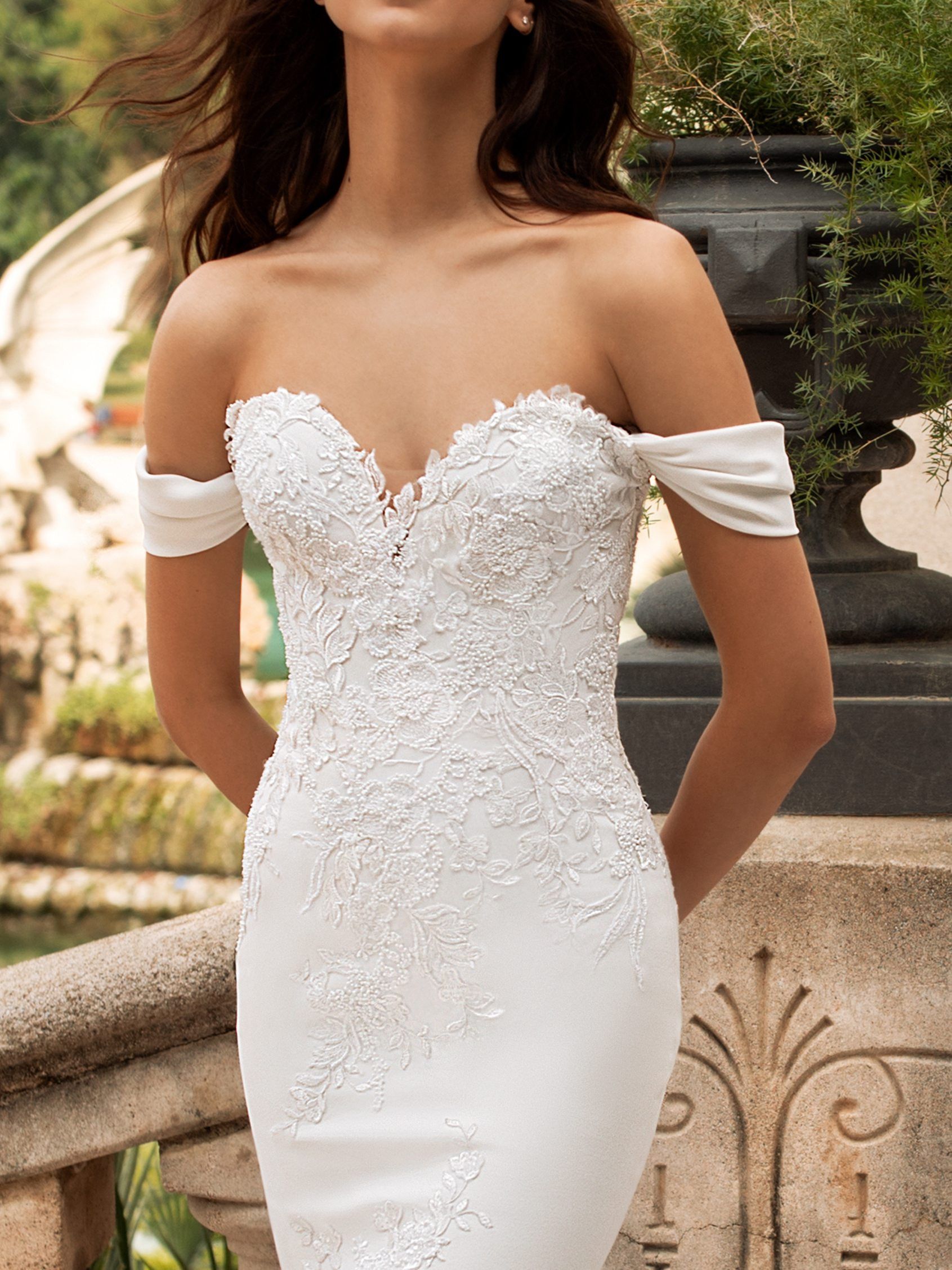 A person wearing a white, off-the-shoulder wedding dress with intricate floral lace bodice detailing outdoors.