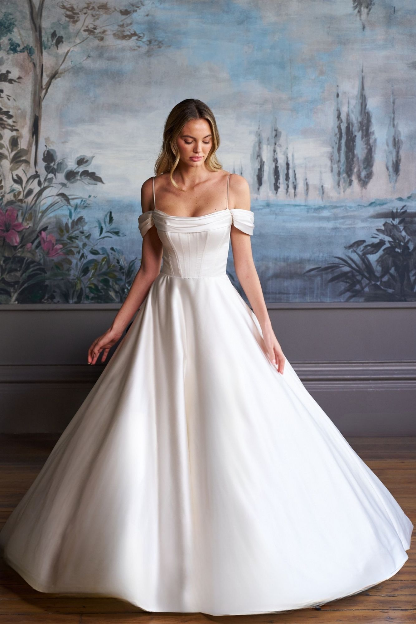 A person wearing a white, off-the-shoulder A-line wedding gown with a satin bodice against a floral mural backdrop.