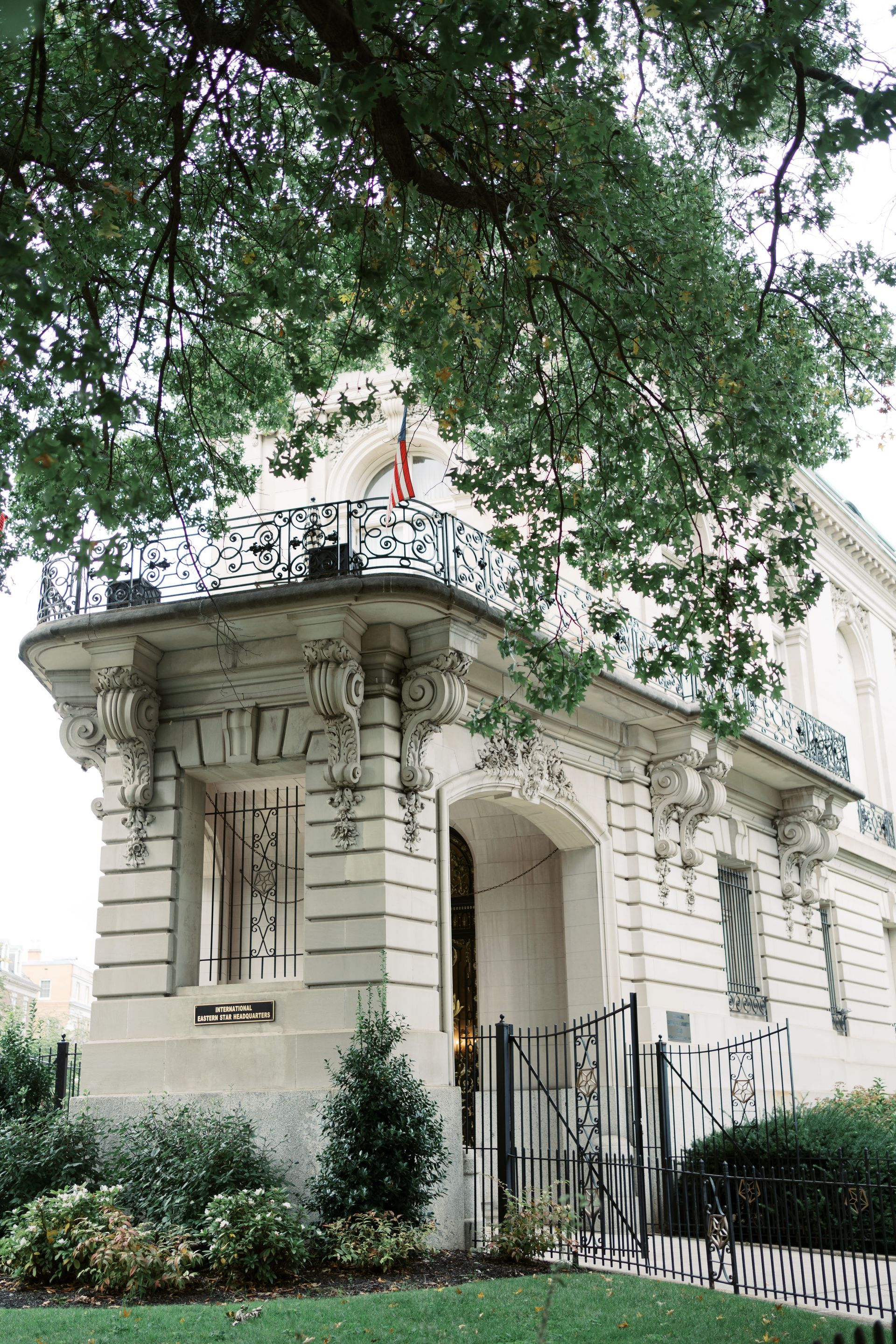 A historic stone mansion with an ornate balcony and iron gate, partially shaded by the canopy of a large tree.