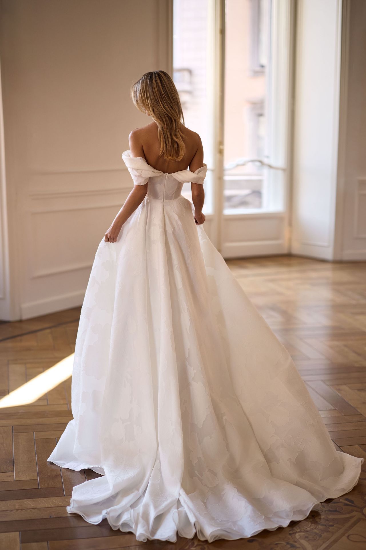 A person viewed from behind in a white, off-the-shoulder wedding gown with a full train in a sunlit, elegant room.