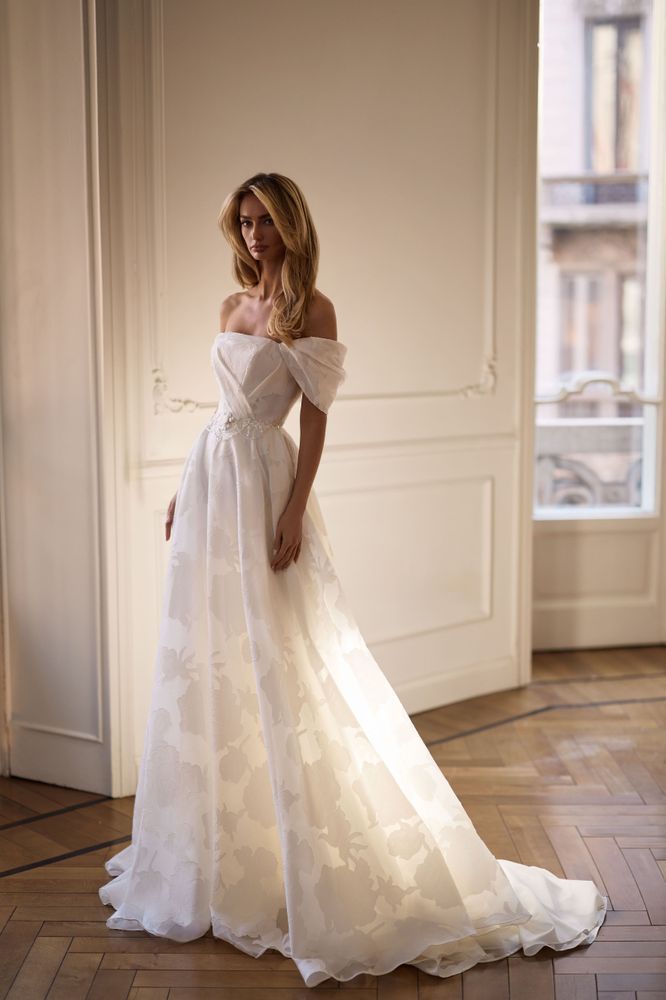 A model wearing a white, off-the-shoulder wedding gown with a full skirt, standing in a sunlit room.