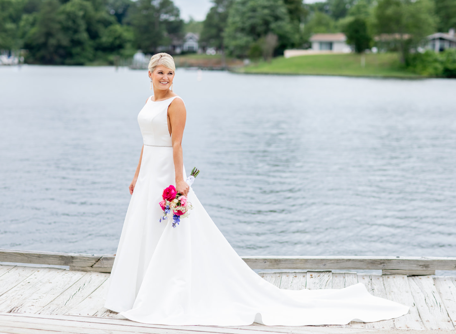 A person in a white wedding dress holds a colorful bouquet, posing on a wooden dock by a calm lake.