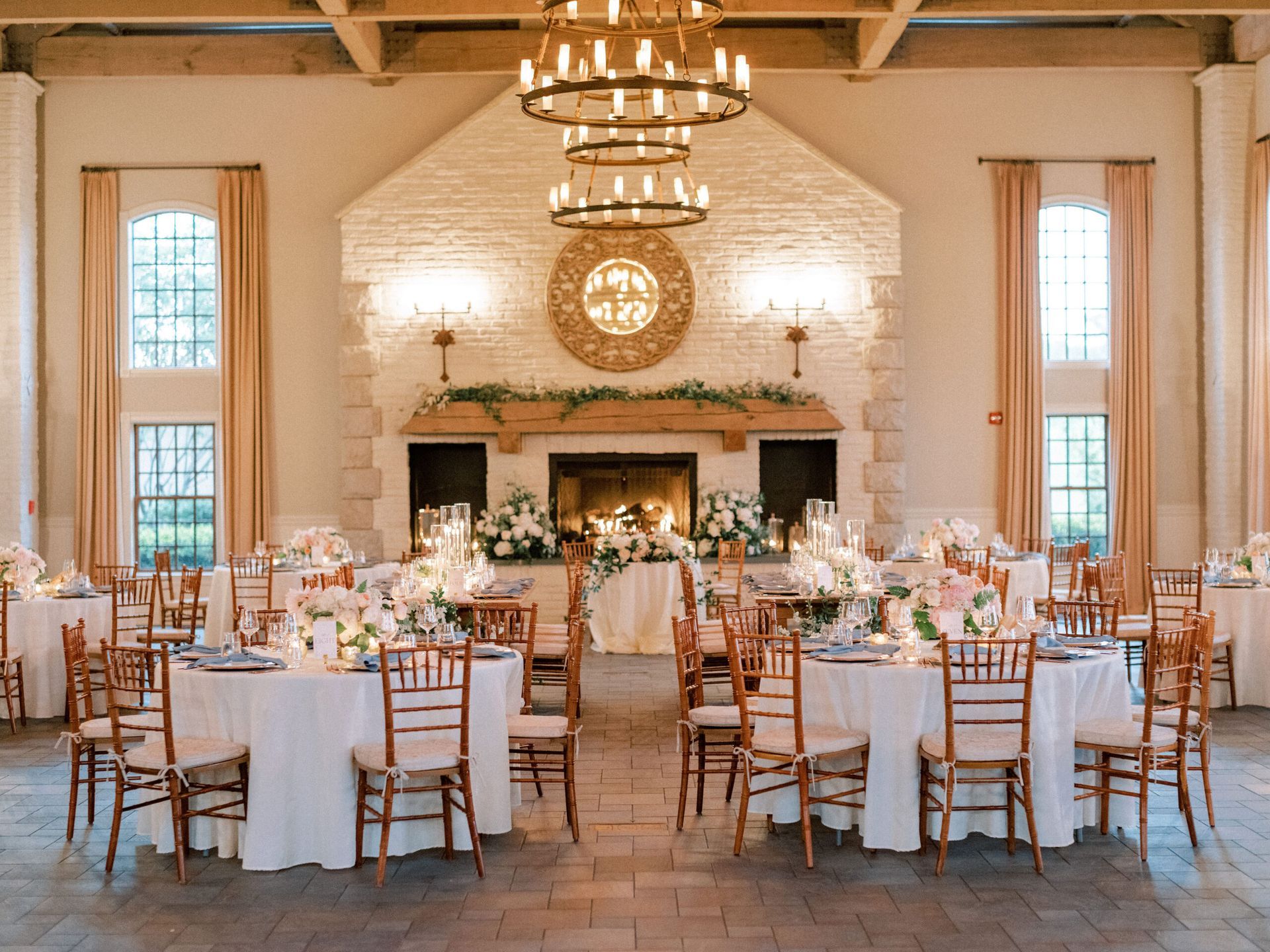 A wedding reception venue with round tables, white tablecloths, and wooden chairs, featuring a large stone fireplace.